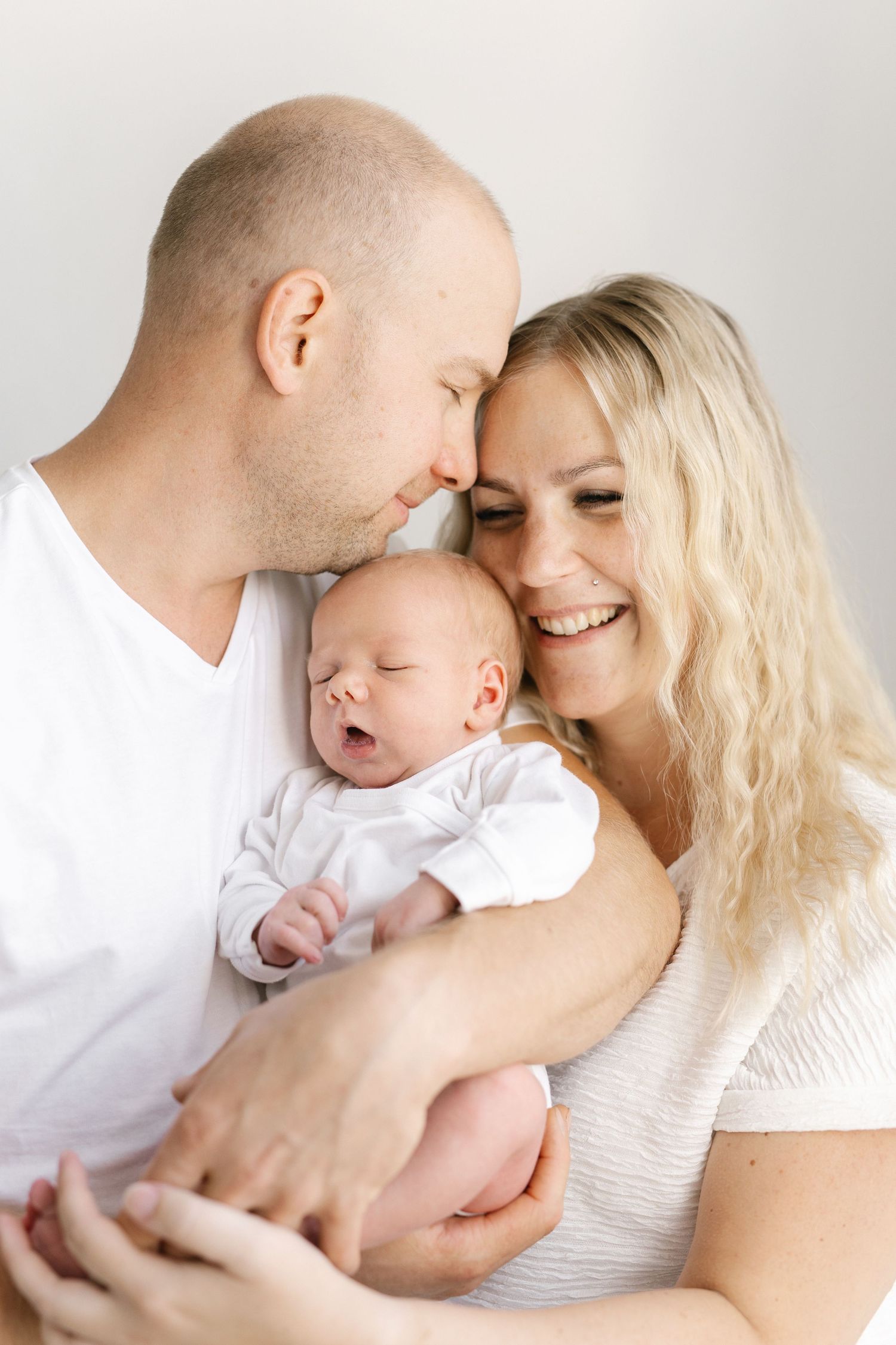 Happy parents smiling and holding their sleeping newborn baby dressed in white against a light background.