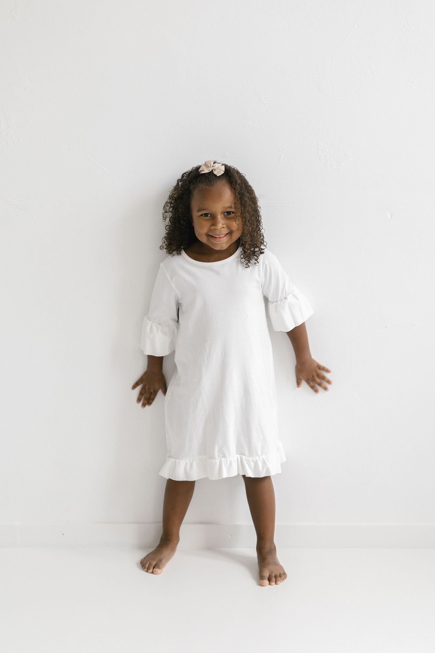 Young girl in white ruffle dress with bell sleeves, smiling and posing barefoot against white background.