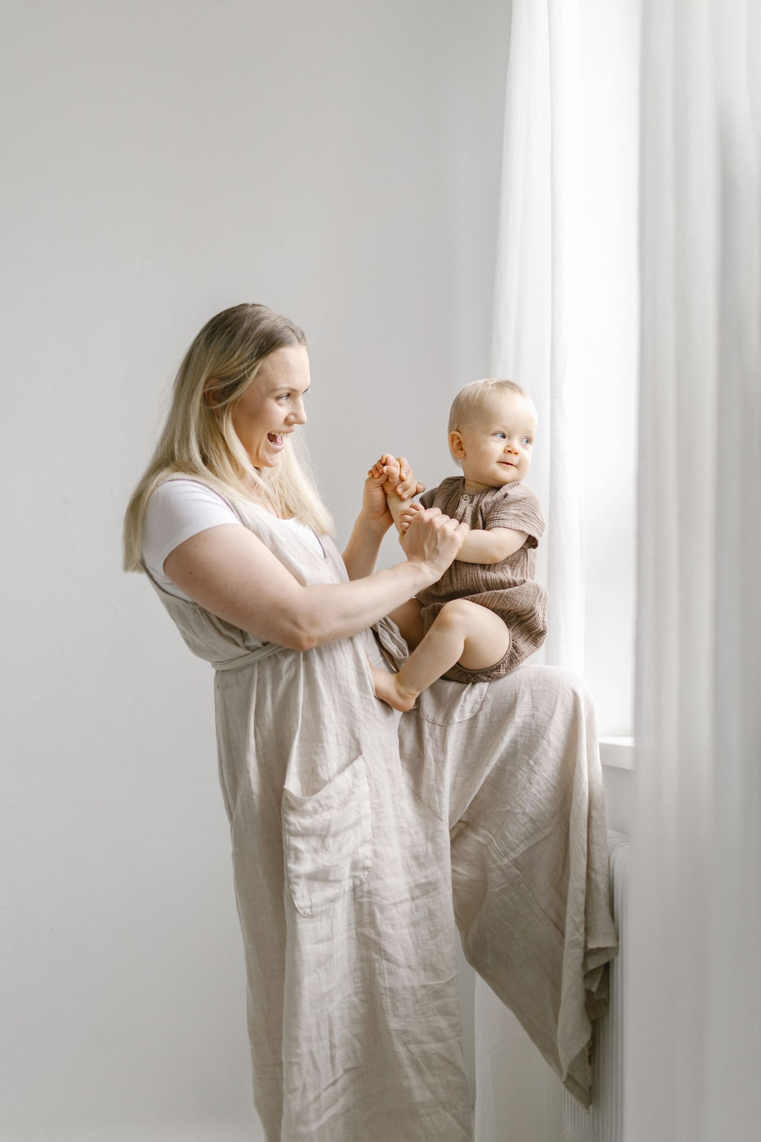 A smiling mother in a linen dress holds her baby boy near a bright window in a minimalist white room.