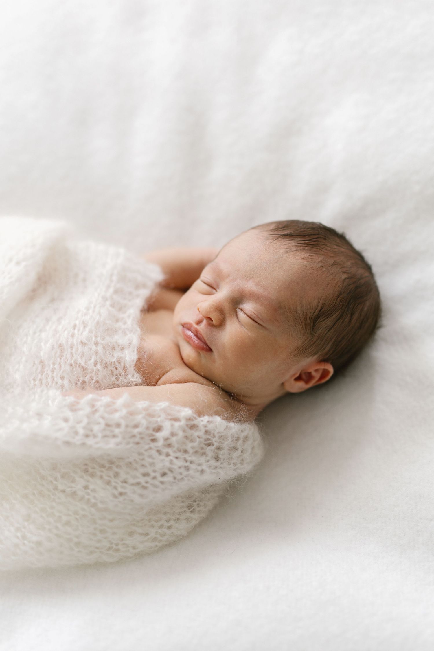 Peaceful newborn baby sleeping wrapped in a soft white knit blanket on a white fluffy surface.