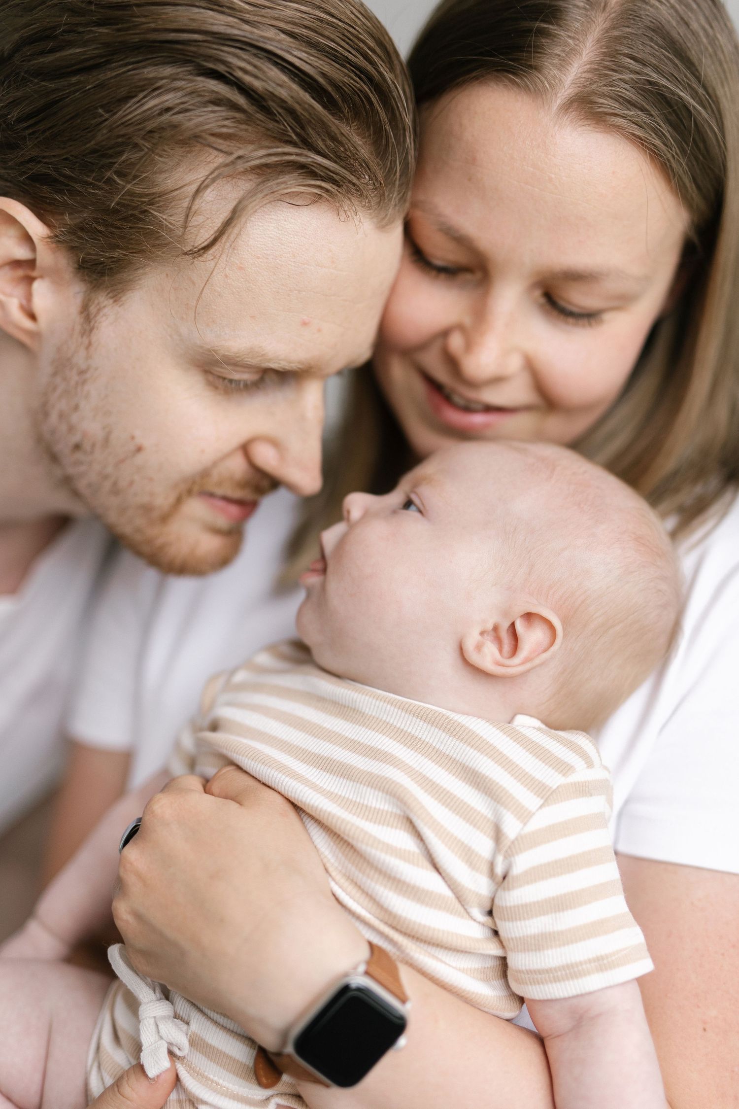 Young parents in white shirts lovingly gaze at their newborn baby in a striped onesie.