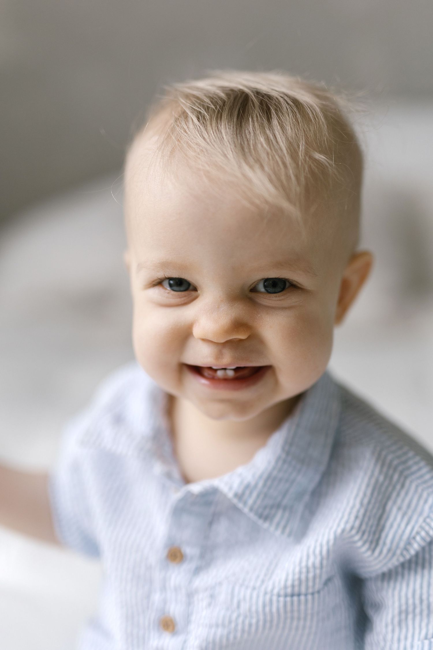 Smiling baby boy with blue eyes and blonde hair wearing a light blue striped shirt, looking at the camera.