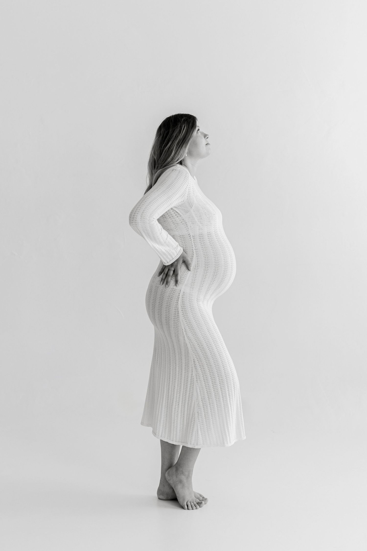 Black and white maternity photo of pregnant woman in white ribbed dress, side profile pose against light background.