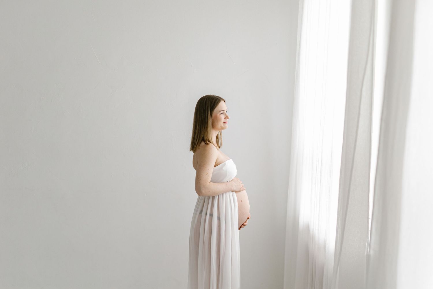 Pregnant woman in a flowing white maternity gown cradling her belly beside a sheer curtain in a bright, minimalist maternity photoshoot.