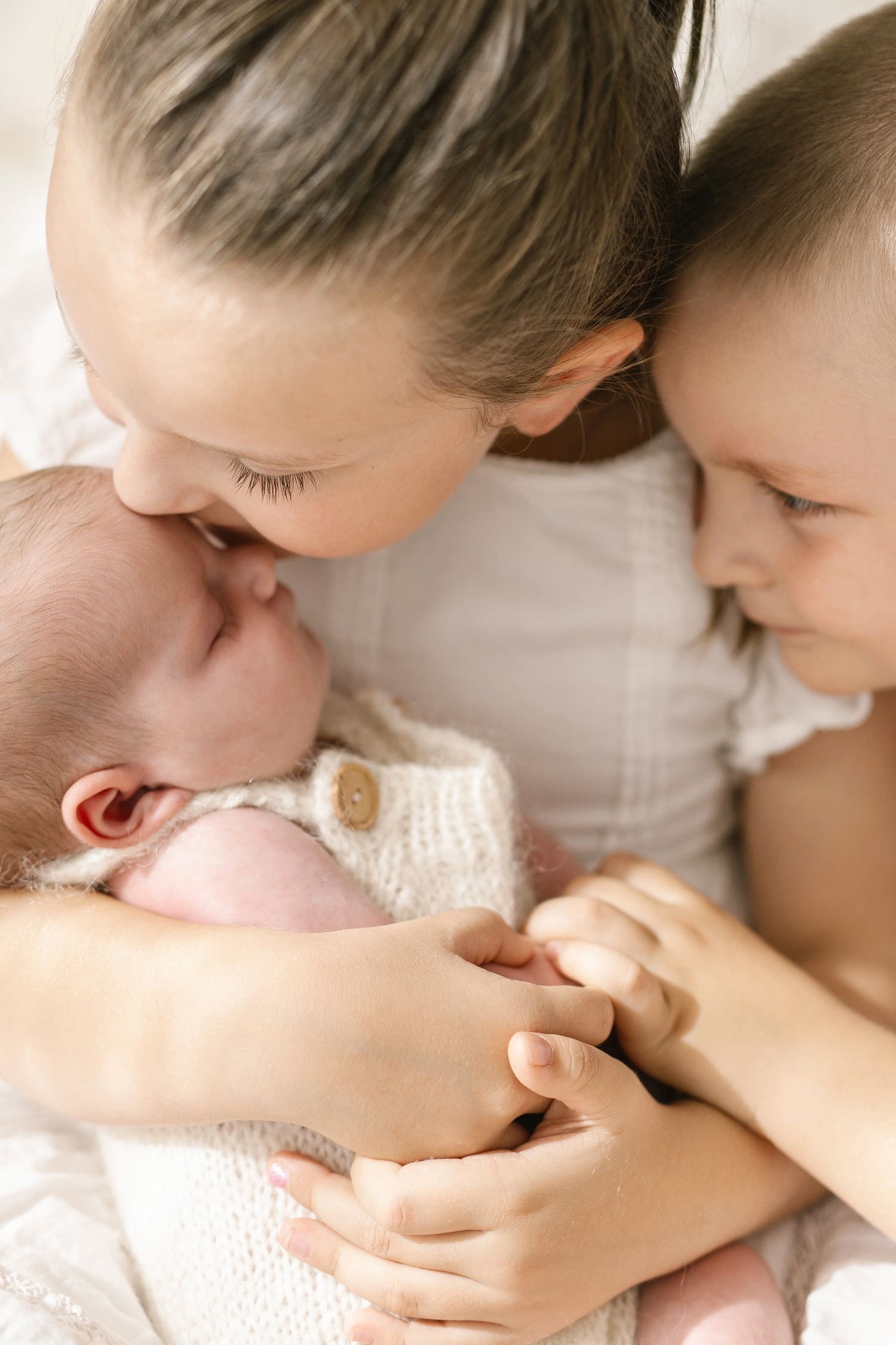 Sister and brother tenderly holding and kissing a sleeping newborn baby wrapped in a white knit blanket.