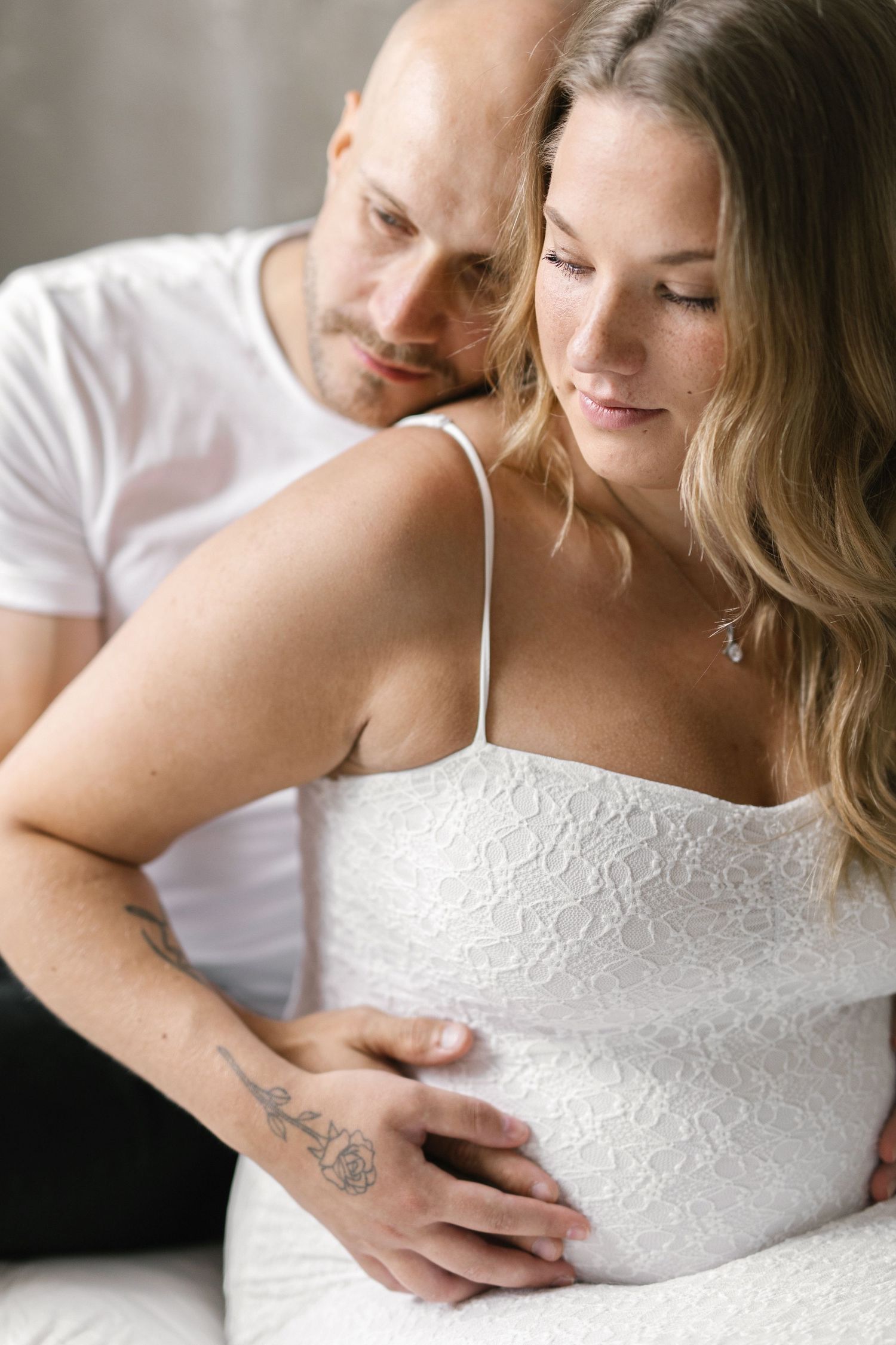 A couple shares a tender moment, man embracing pregnant woman in white lace dress, his tattooed hand resting on her baby bump.
