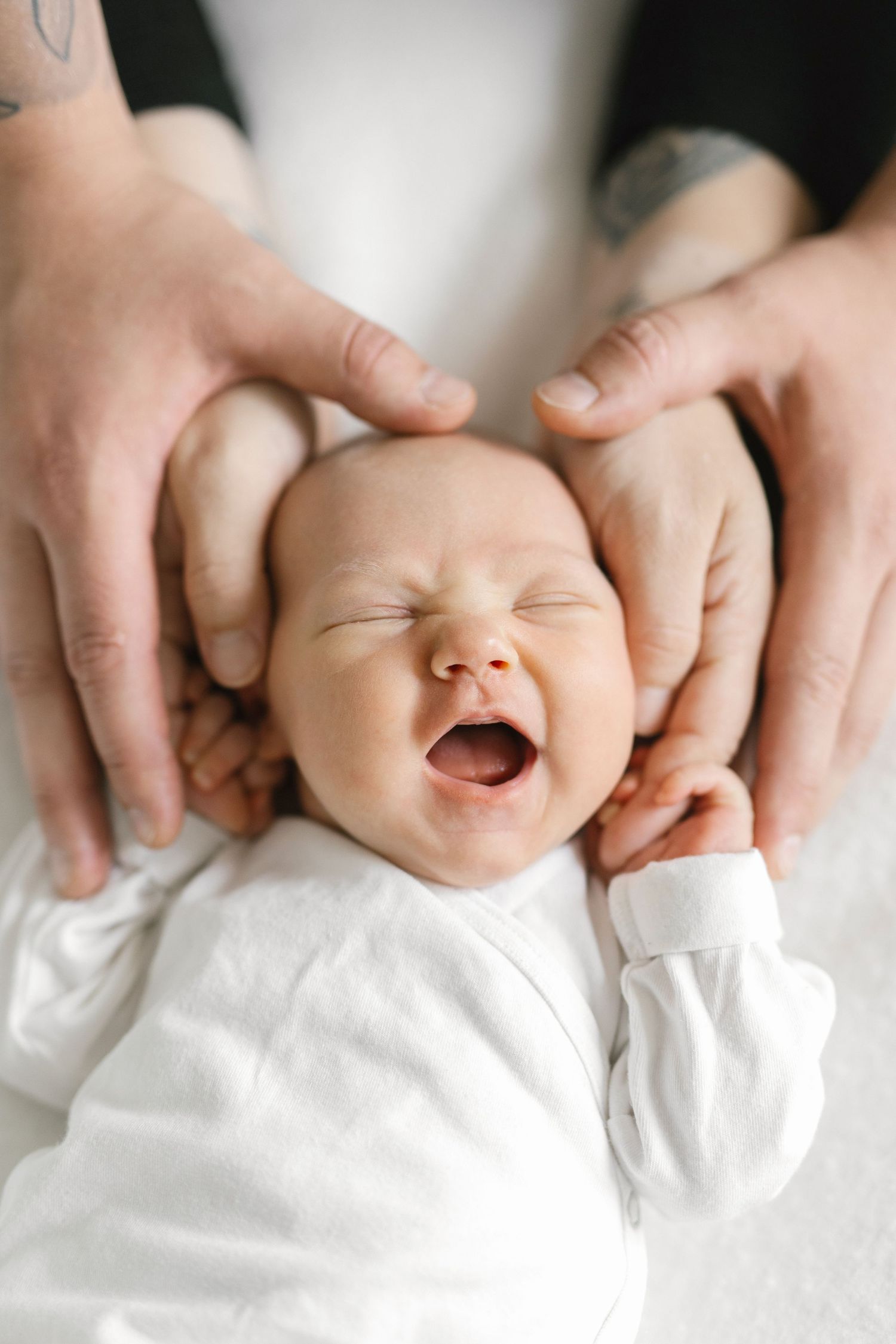 Newborn baby yawning while cradled in parents hands, wearing white onesie.