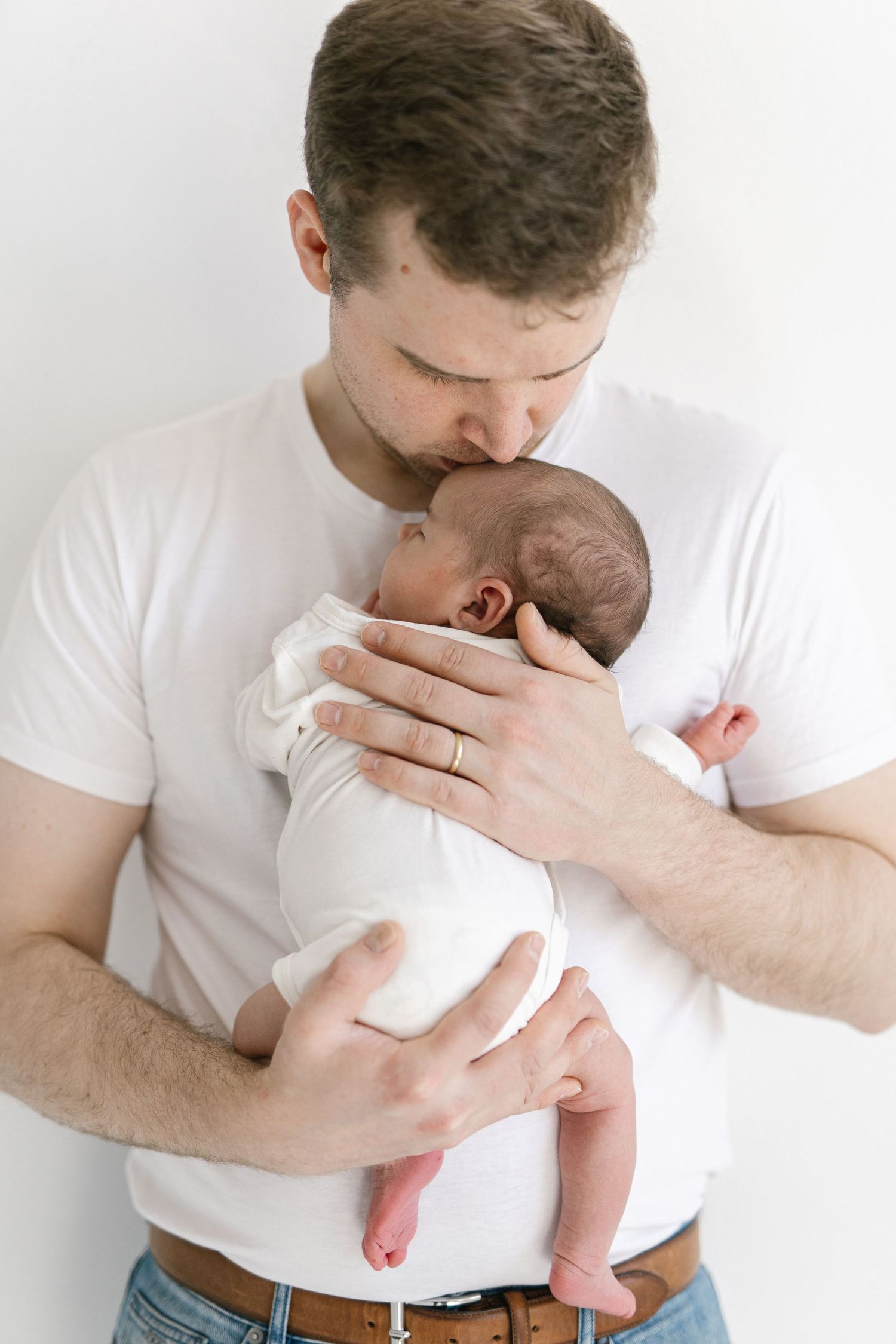 A father in a white t-shirt tenderly kisses his newborn baby while cradling them gently against his chest.