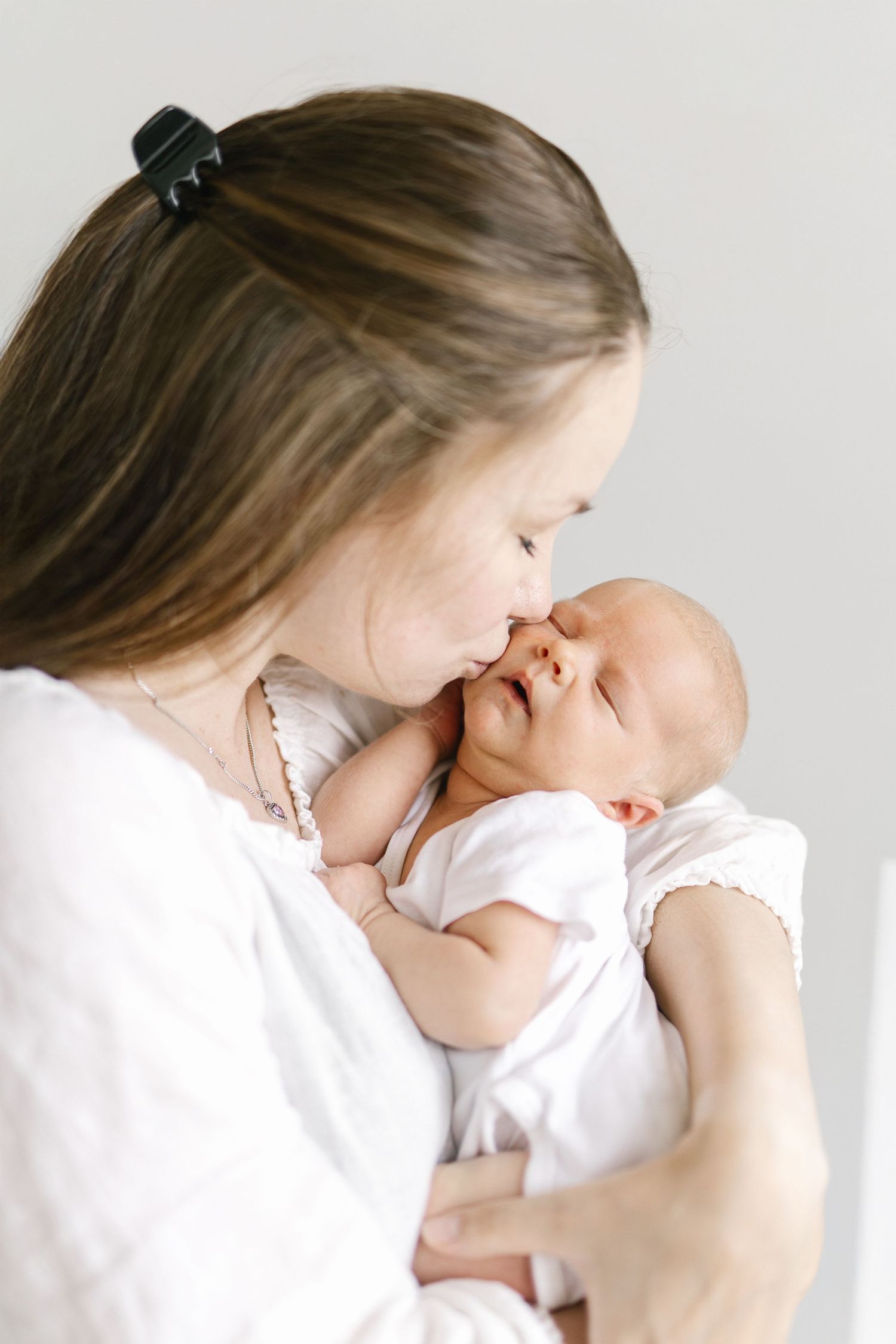 A tender moment as a young mother in white gently kisses her sleeping newborn baby dressed in white.