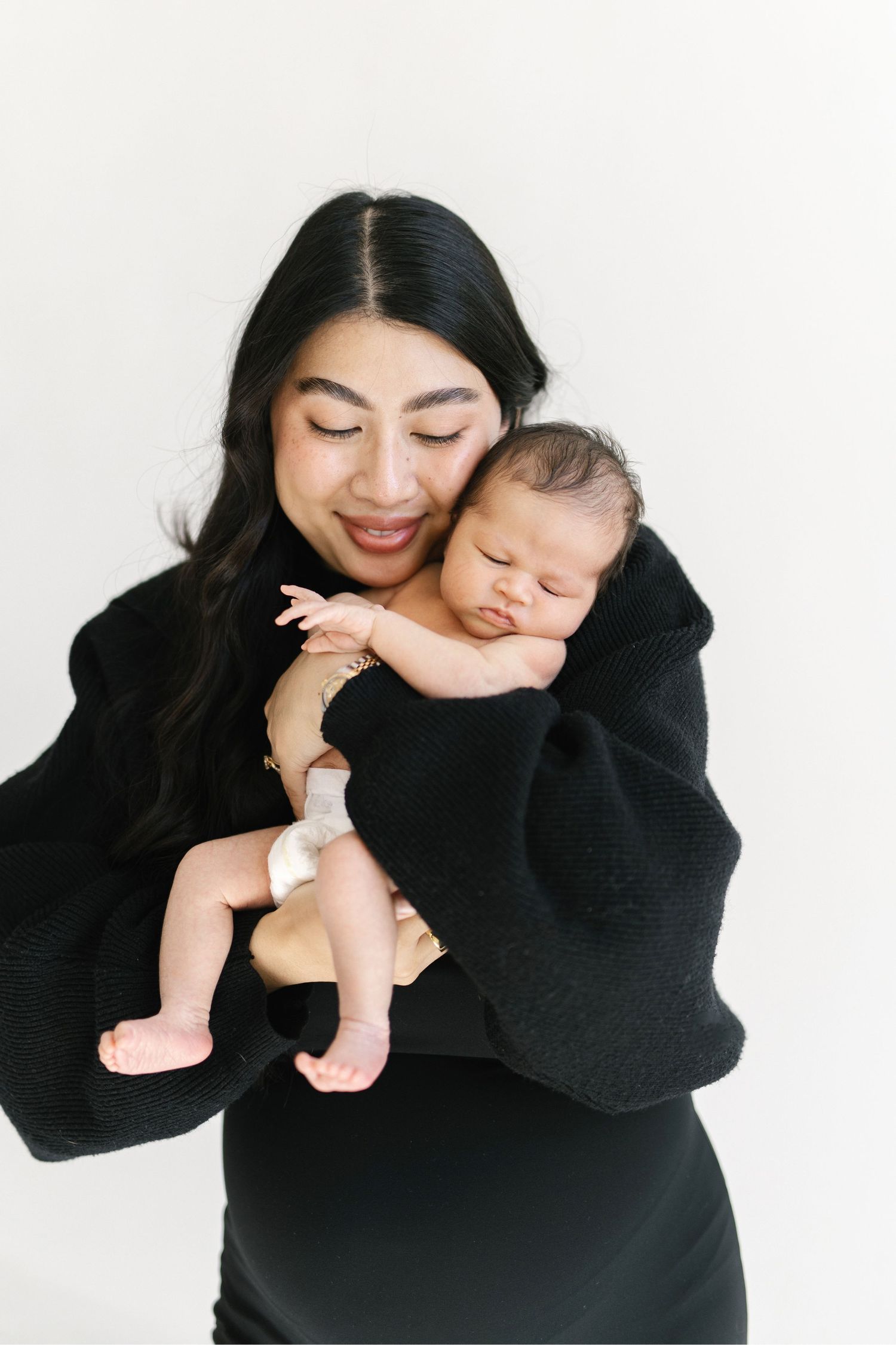 Smiling mother in black sweater gently cradling her sleeping newborn baby against her cheek.