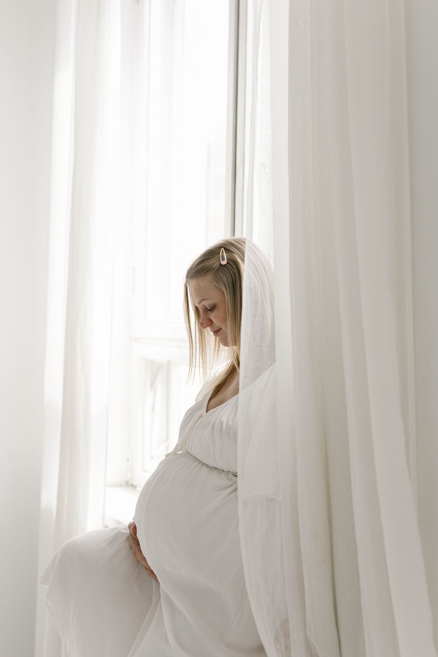 Pregnant woman in white dress holding baby bump by sheer curtains in bright room.