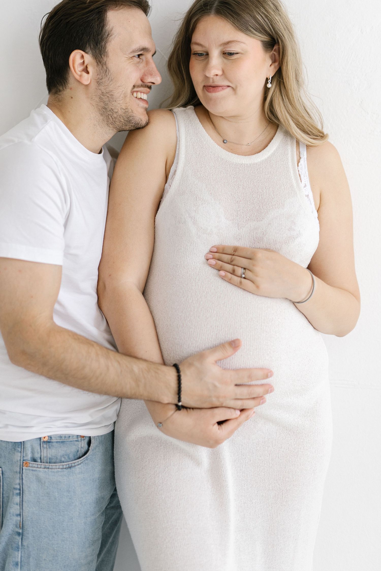 Happy couple embracing, pregnant woman in white dress cradling baby bump with partner smiling beside her against white wall.