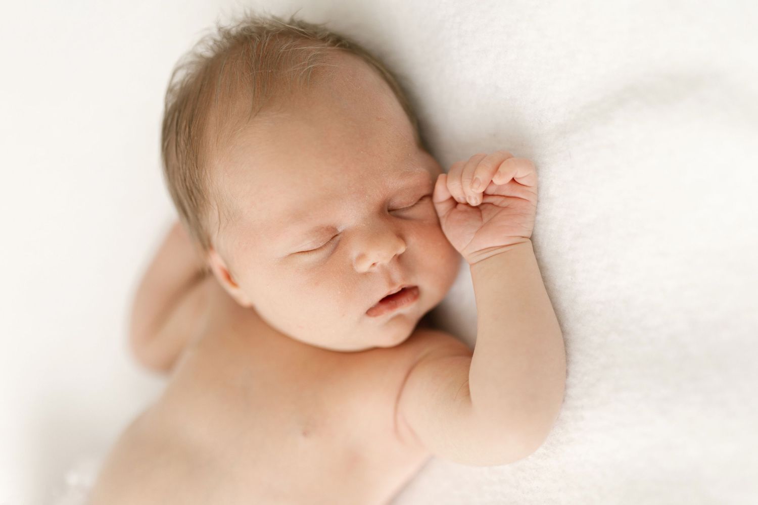 Sleeping newborn baby lying on a soft white blanket with one tiny hand raised near the face in a peaceful newborn portrait.