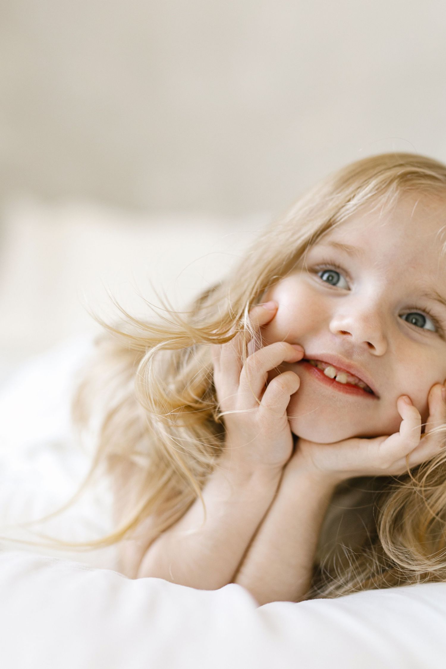 Smiling young girl with long wavy blonde hair and blue eyes rests her chin on her hands while lying on soft white bedding.