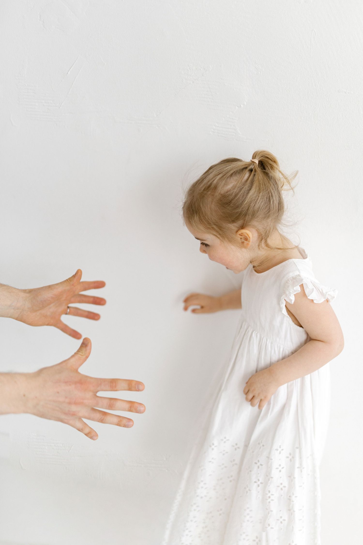 Toddler girl in a white eyelet dress with a hair bun looks down as two adult hands reach toward her against a white wall.
