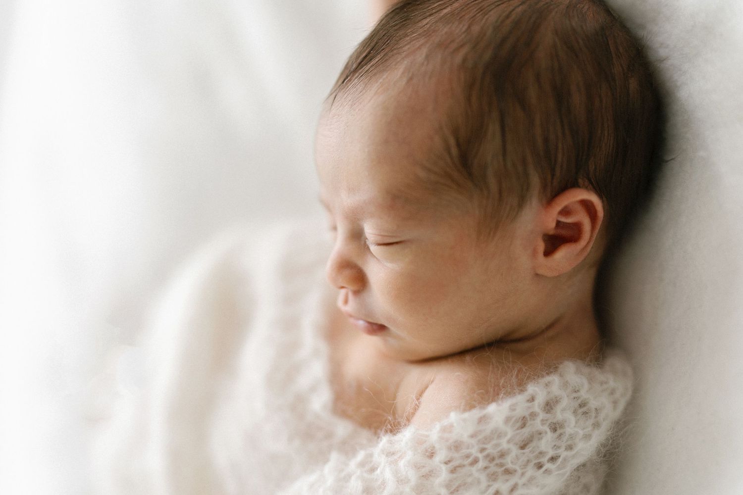 Peaceful newborn baby sleeping wrapped in soft white knit blanket, close-up portrait photography.