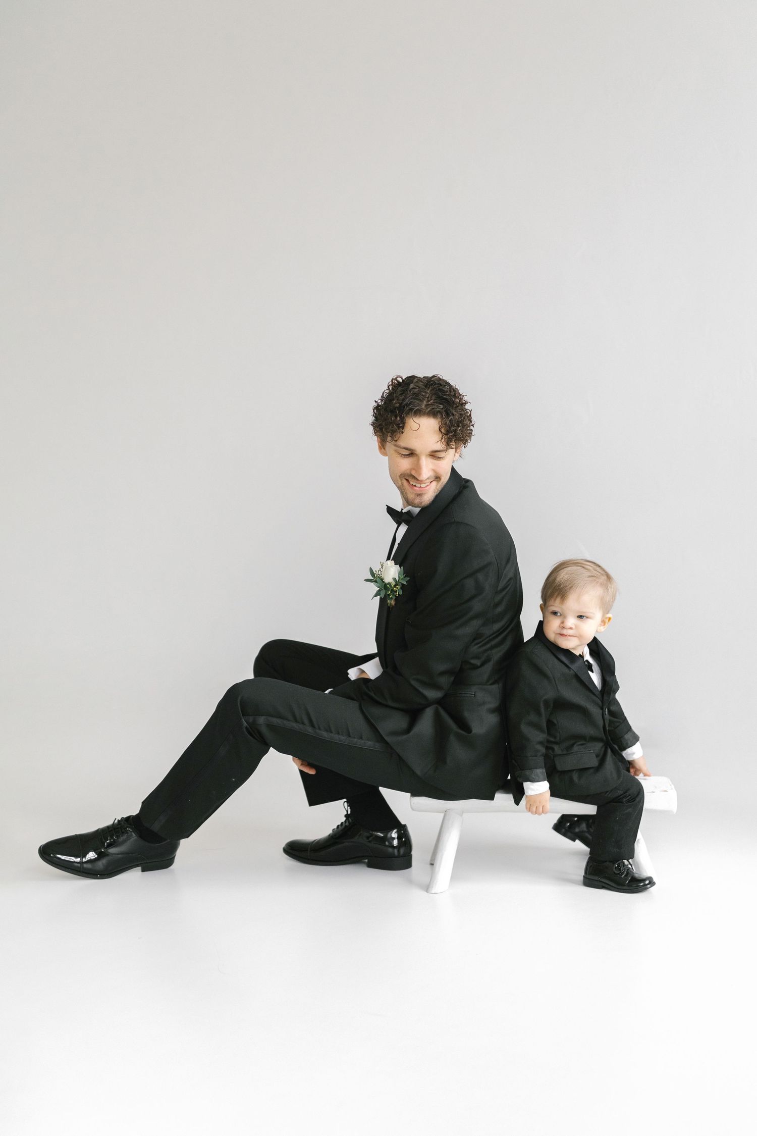 Playful wedding day portrait of two people in matching black suits sitting on white bench.