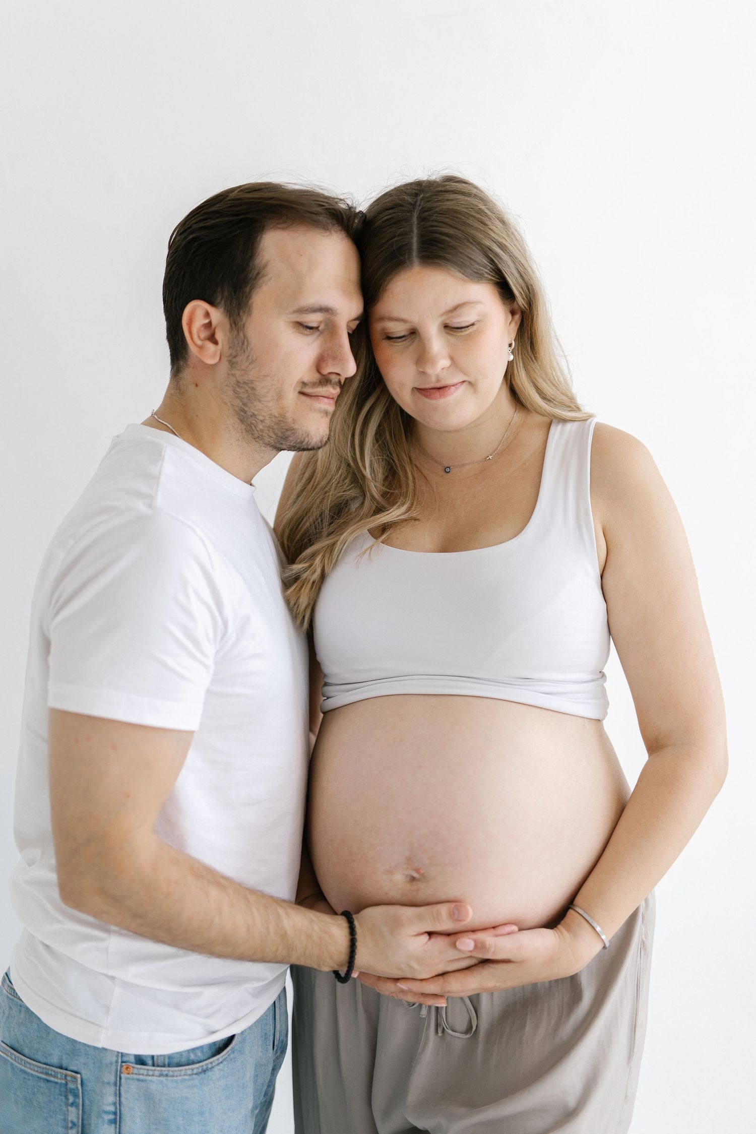 Couple posing for maternity photo, man in white t-shirt embracing pregnant woman holding her bare baby bump.