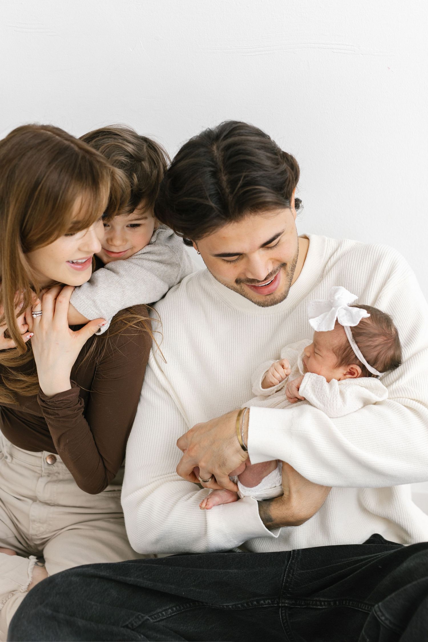 Happy family with parents, toddler, and newborn baby girl in white bow posing together on white background.