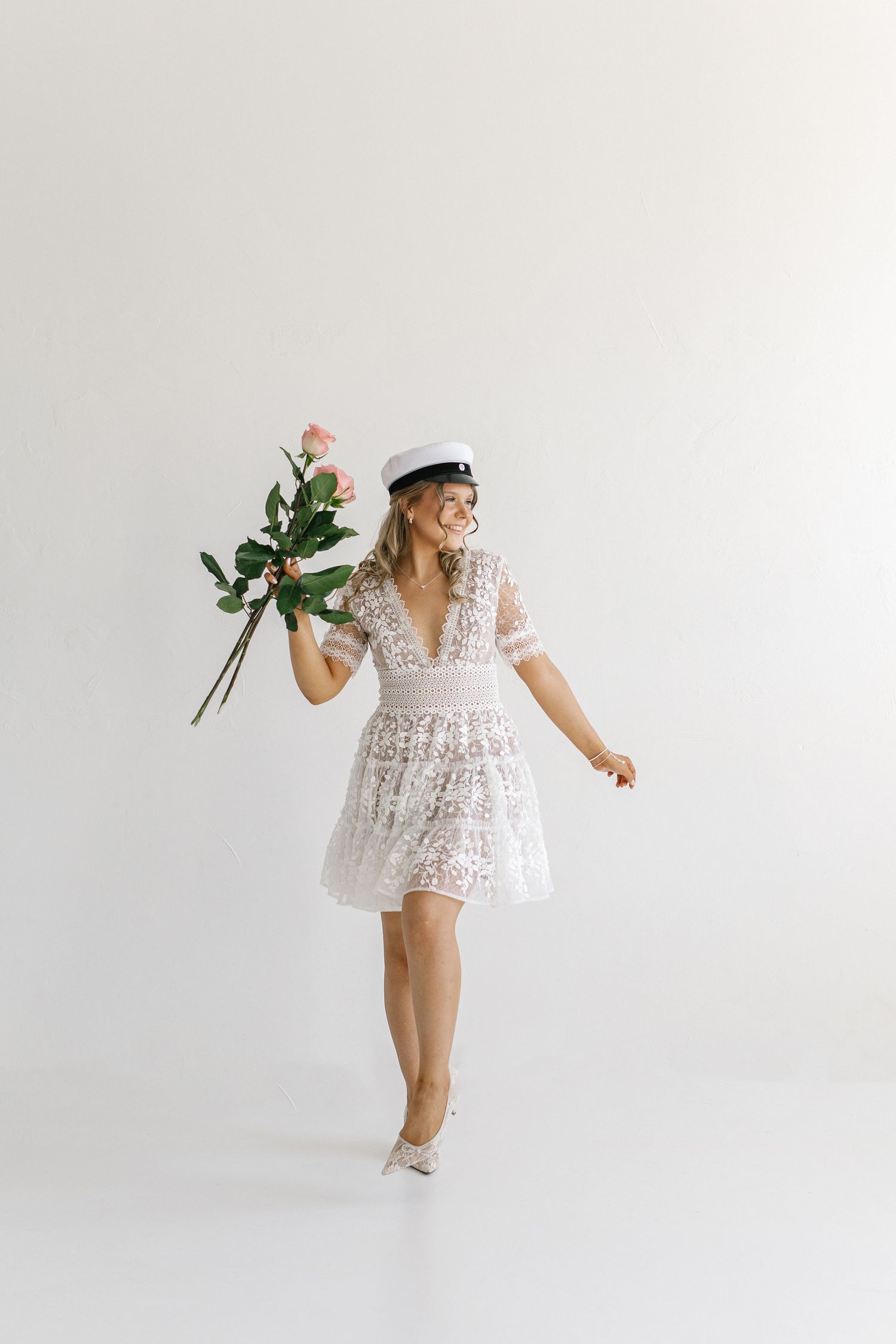 Woman in white lace dress and sailor hat dancing joyfully while holding pink roses bouquet.