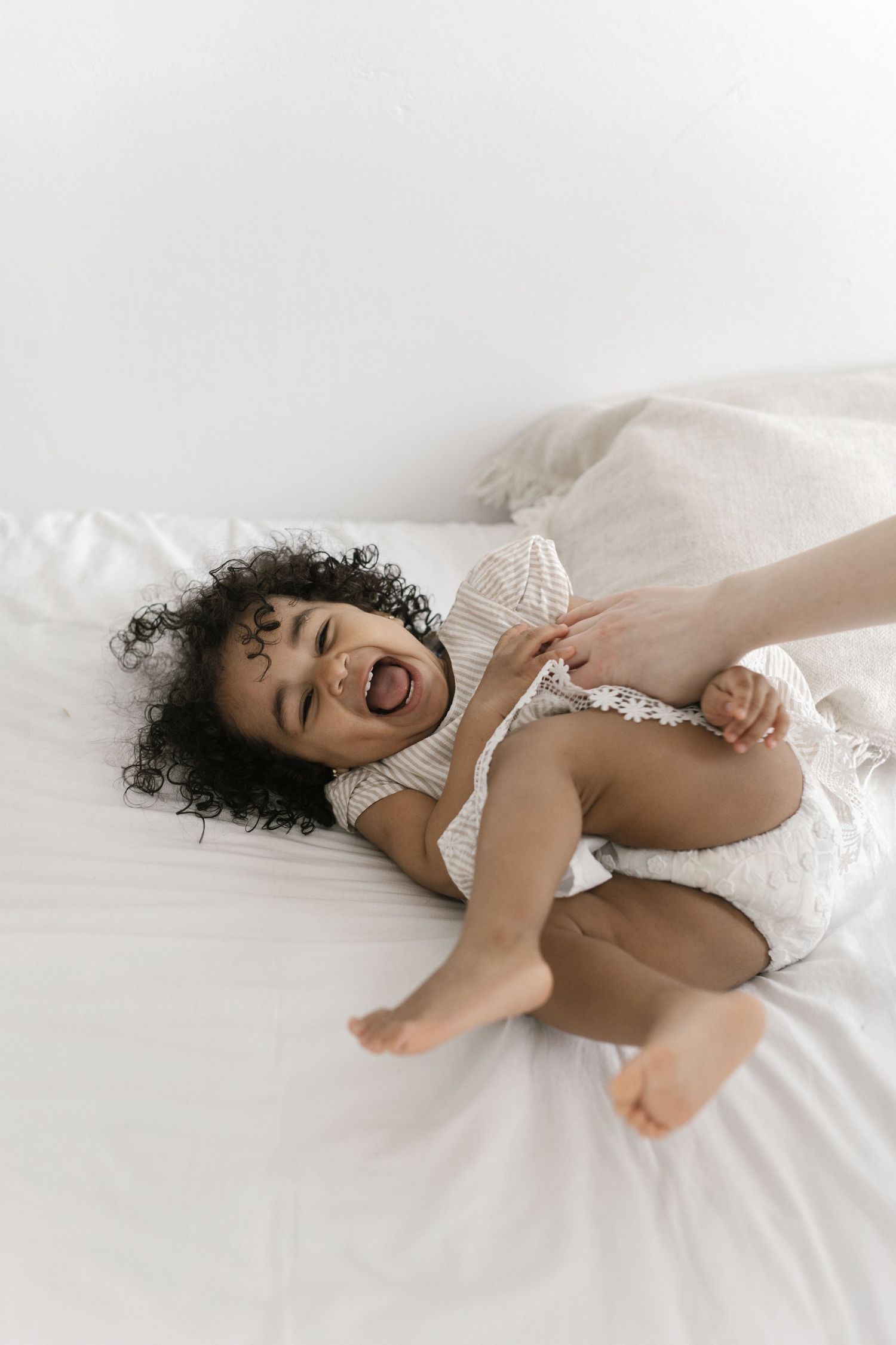 Laughing toddler with curly hair lying on white bed being tickled, wearing a white outfit.