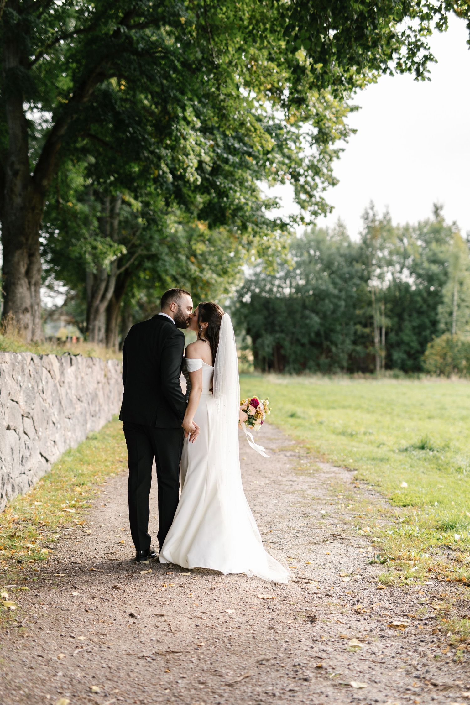A bride and groom share a kiss on a tree-lined gravel path, the bride holding a bouquet in a white gown with veil.