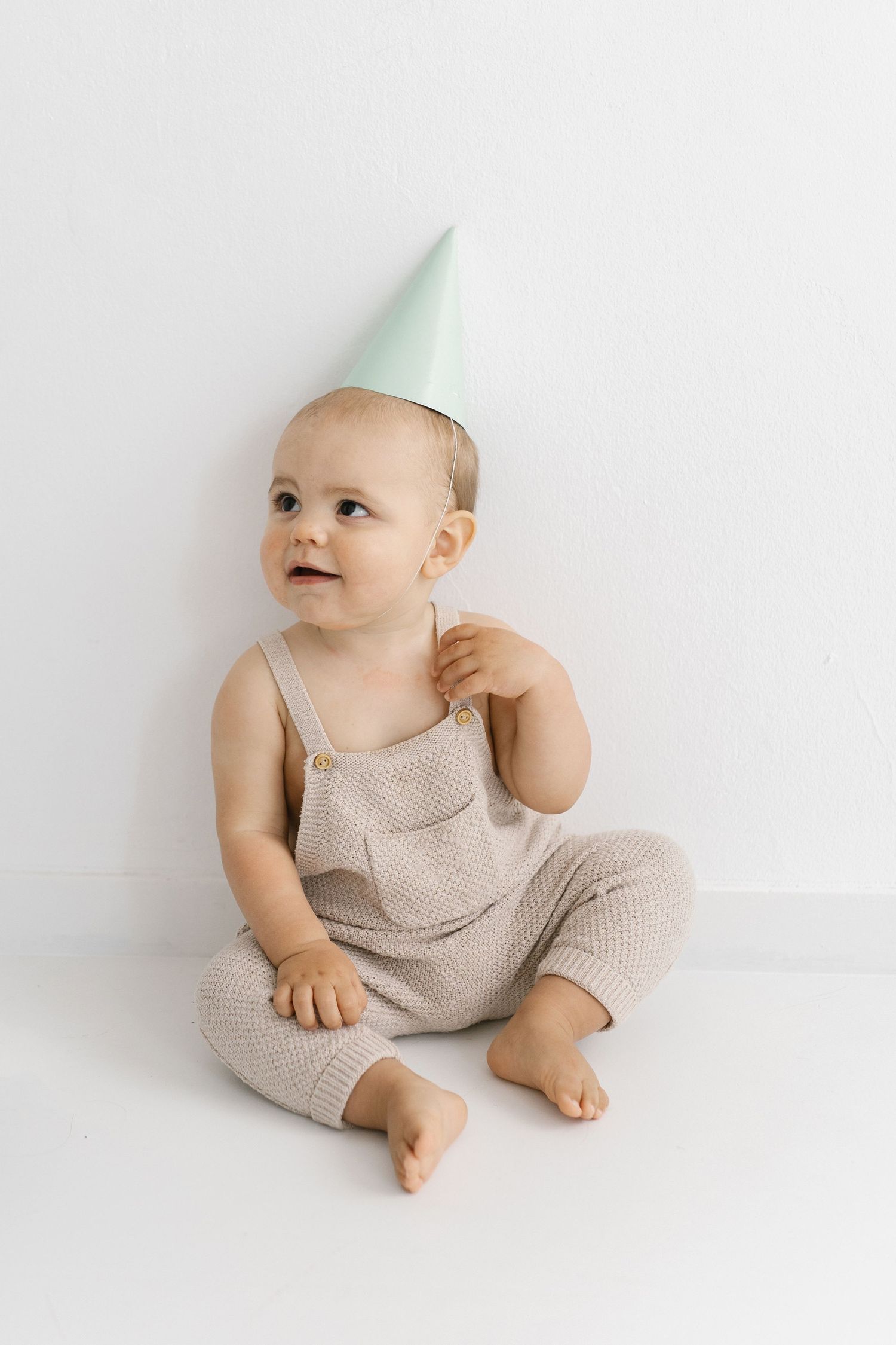 Baby in beige romper wearing mint party hat sits against white background.