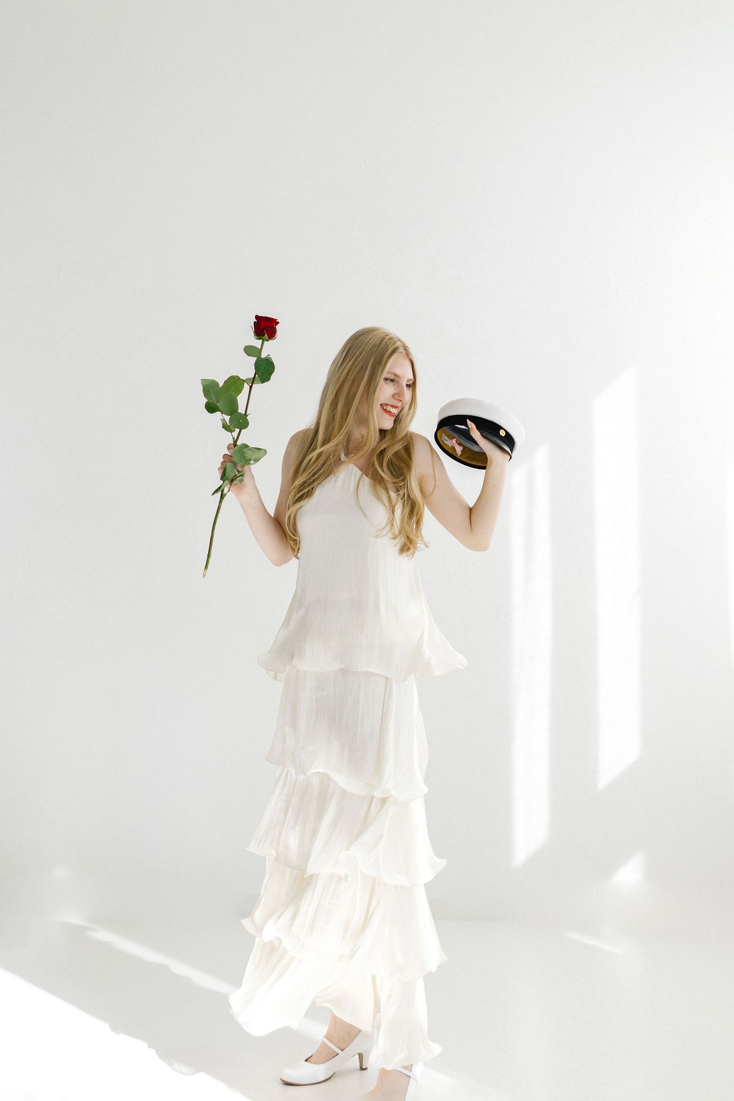 Young woman in white ruffled dress holding a red rose and black hat, posing gracefully in a bright white studio.