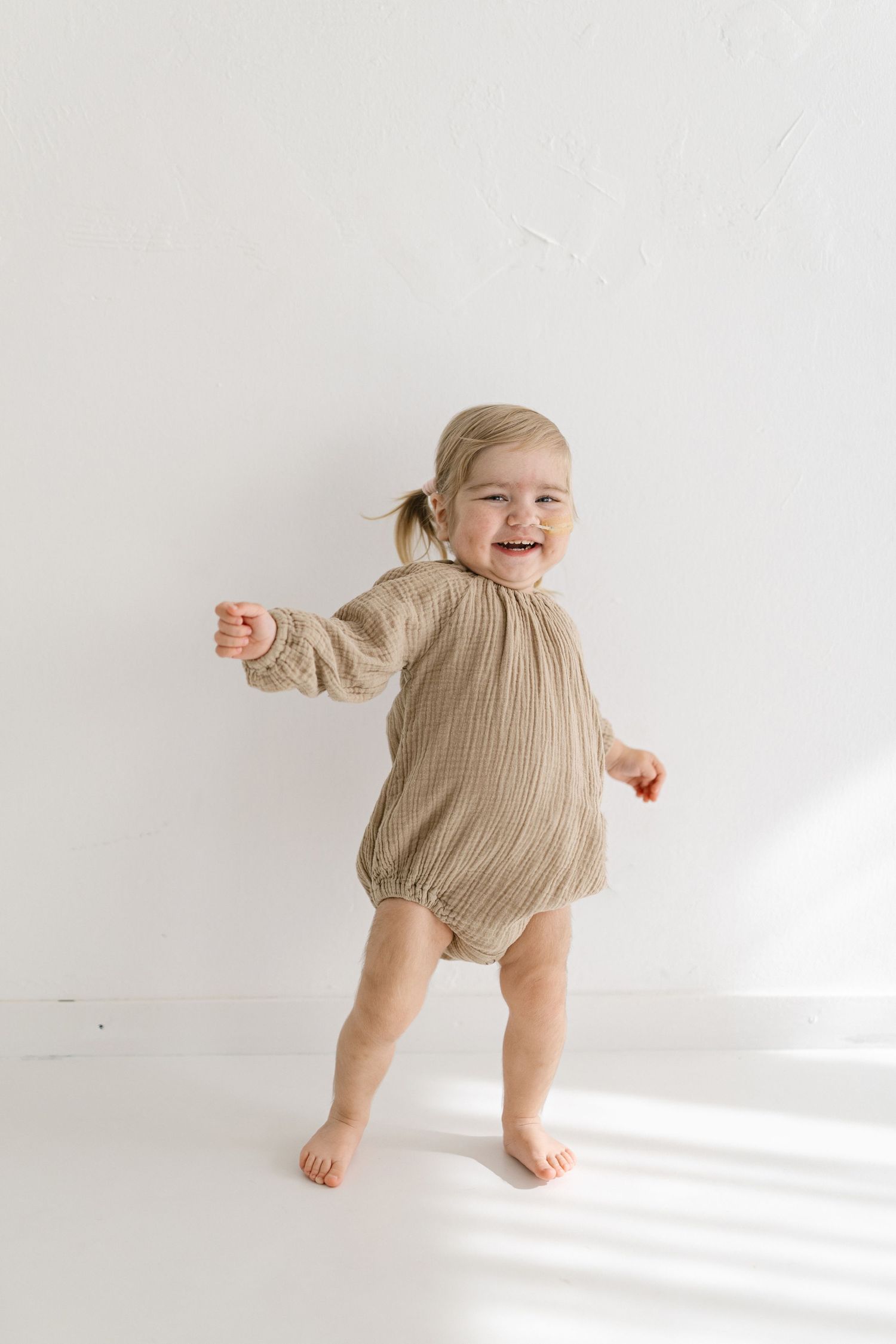 Baby in tan romper walking and laughing against white background.