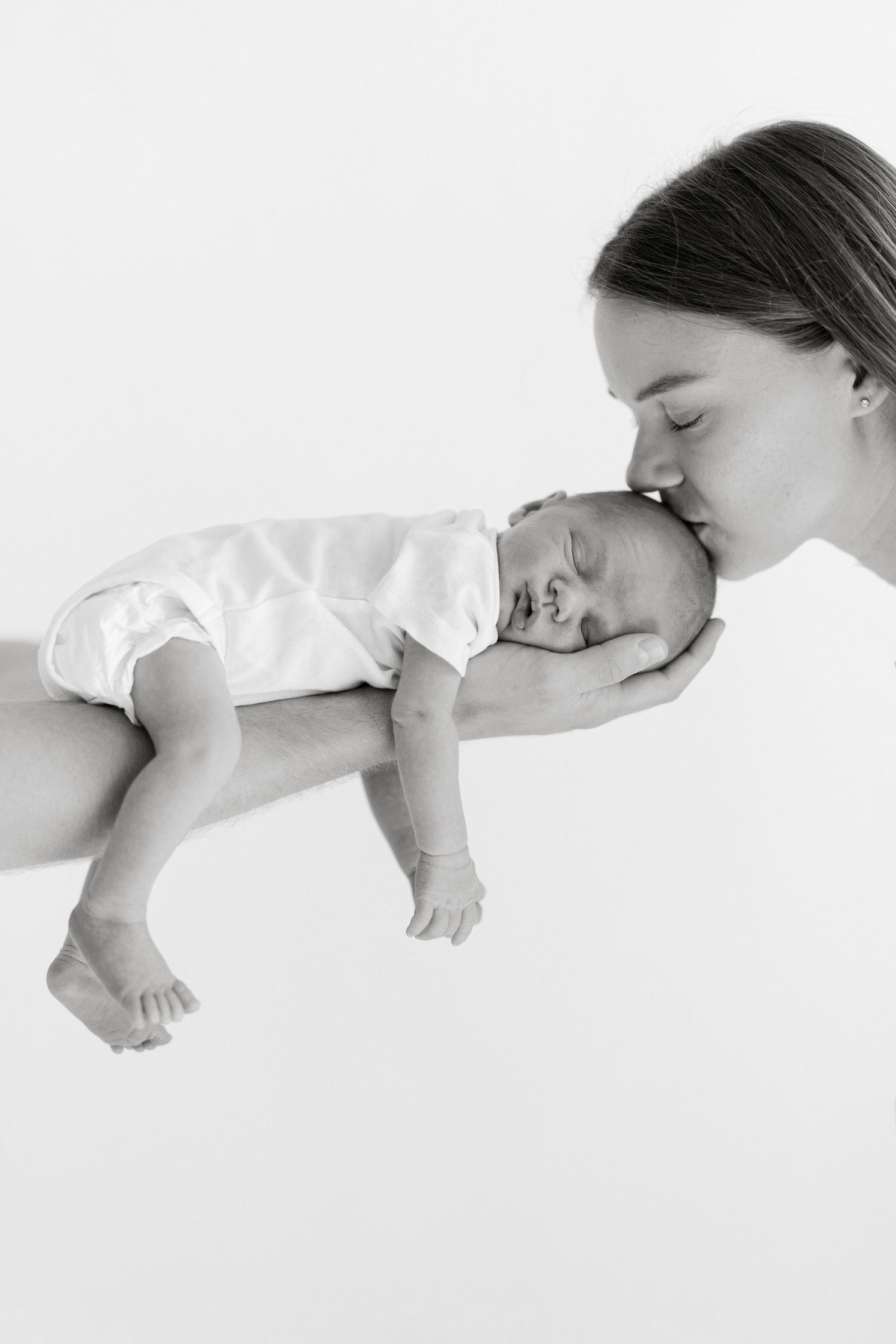 Black and white photo of a mother gently kissing her sleeping newborn baby cradled in her hands.