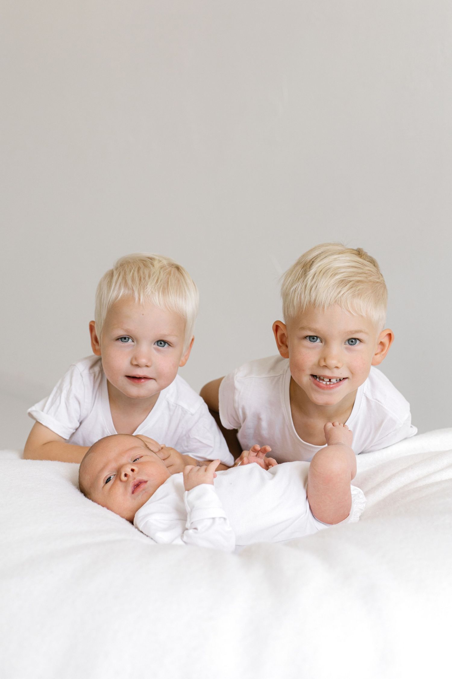 Two blonde toddler boys in white shirts smile while lying on a bed with their newborn baby sibling.