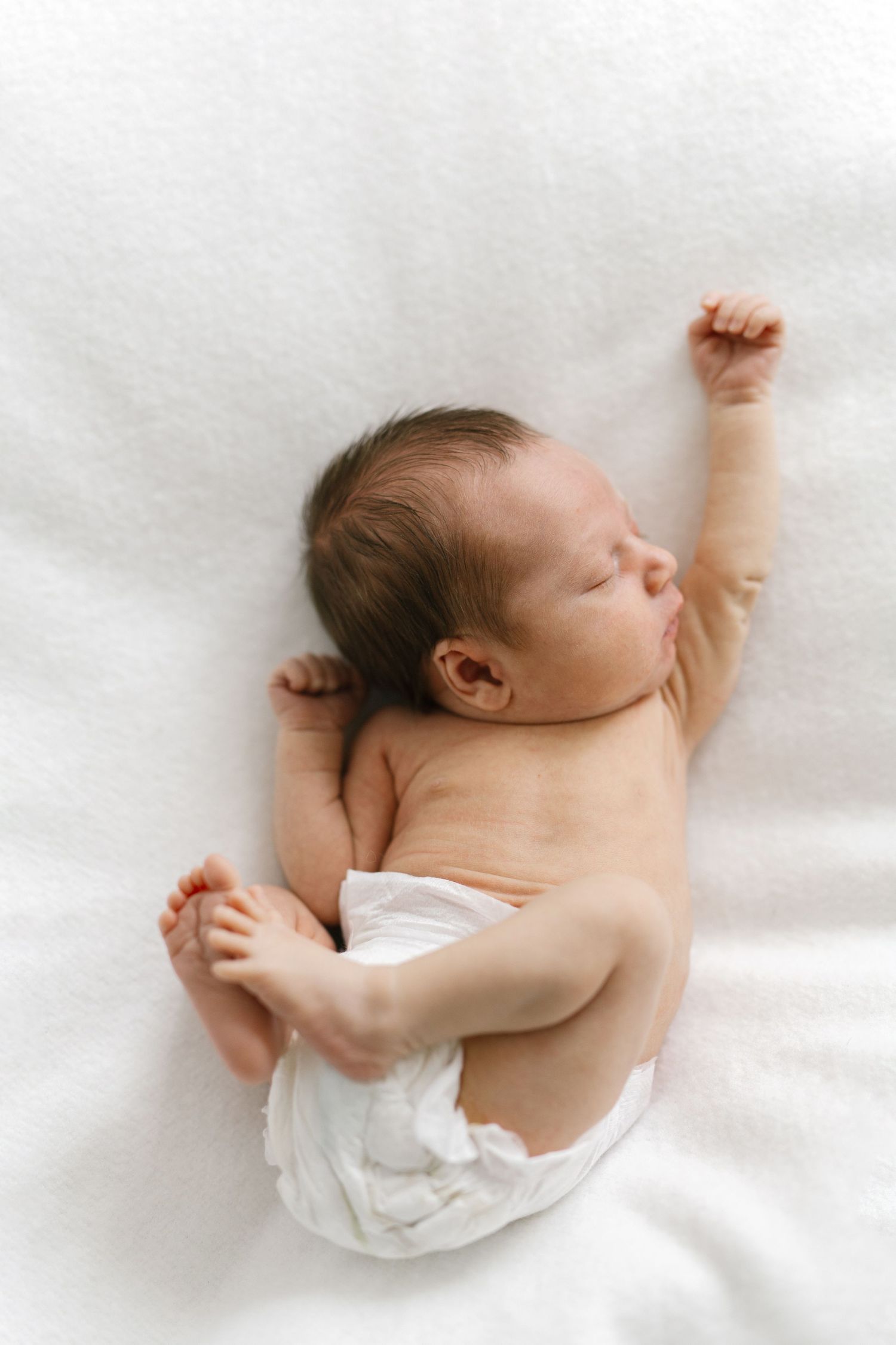 Newborn baby in a white diaper sleeping peacefully on a soft white blanket with one arm raised.