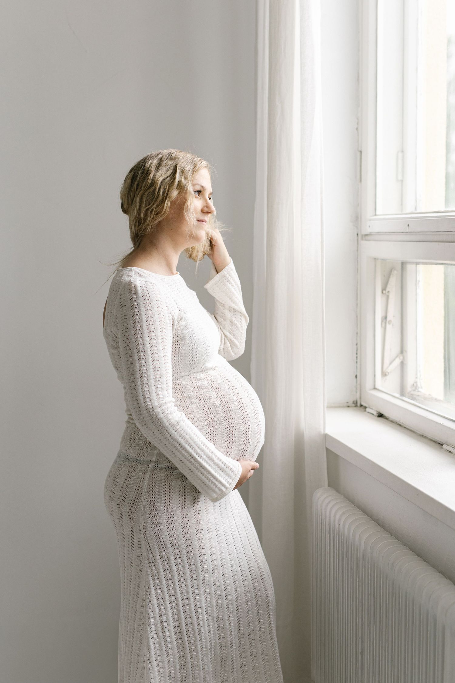 Pregnant woman in white knit dress holding belly, smiling by bright window in serene, light-filled room.
