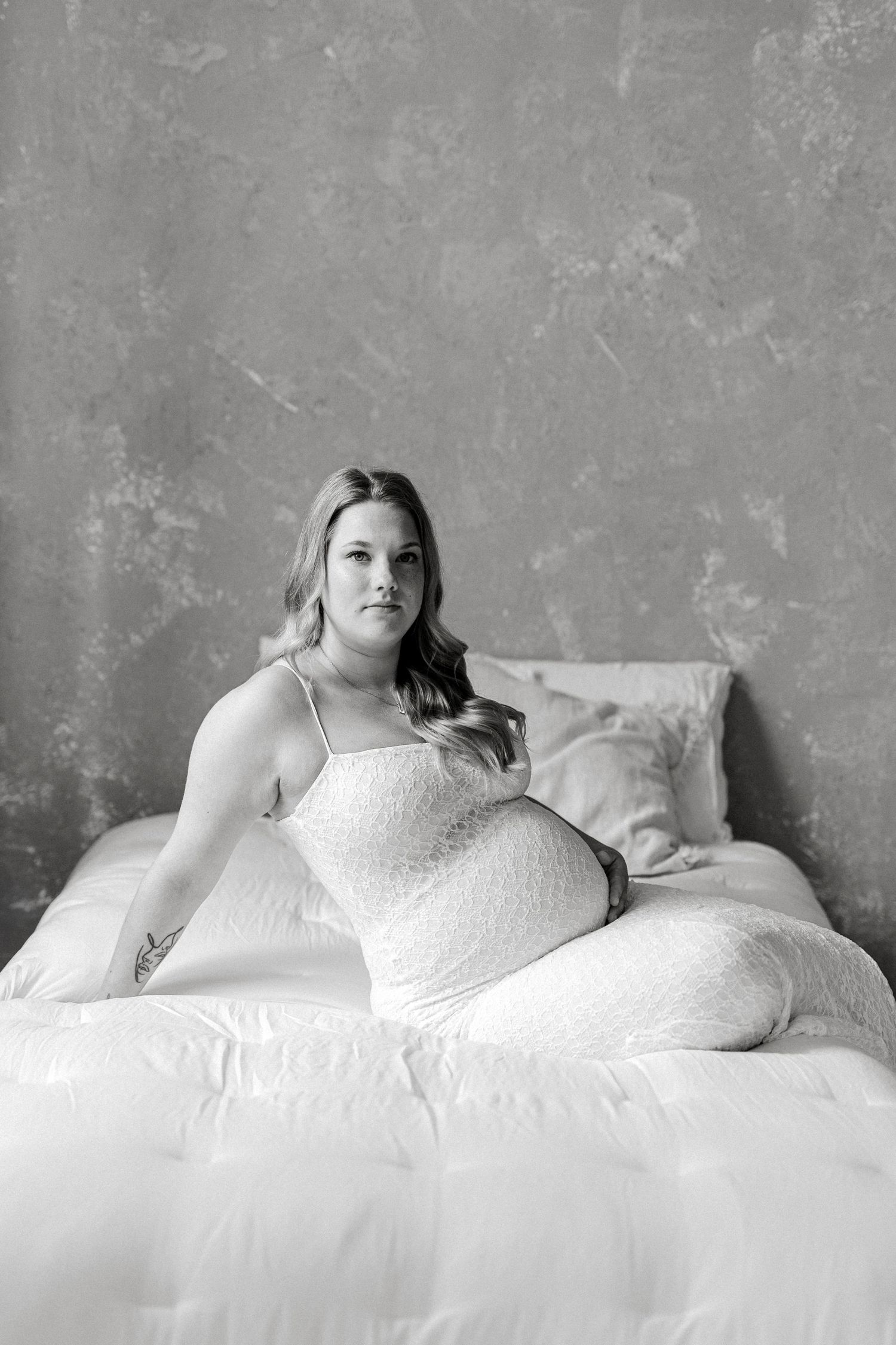 Black and white maternity photo of a pregnant woman in a lace dress sitting on a white bed.
