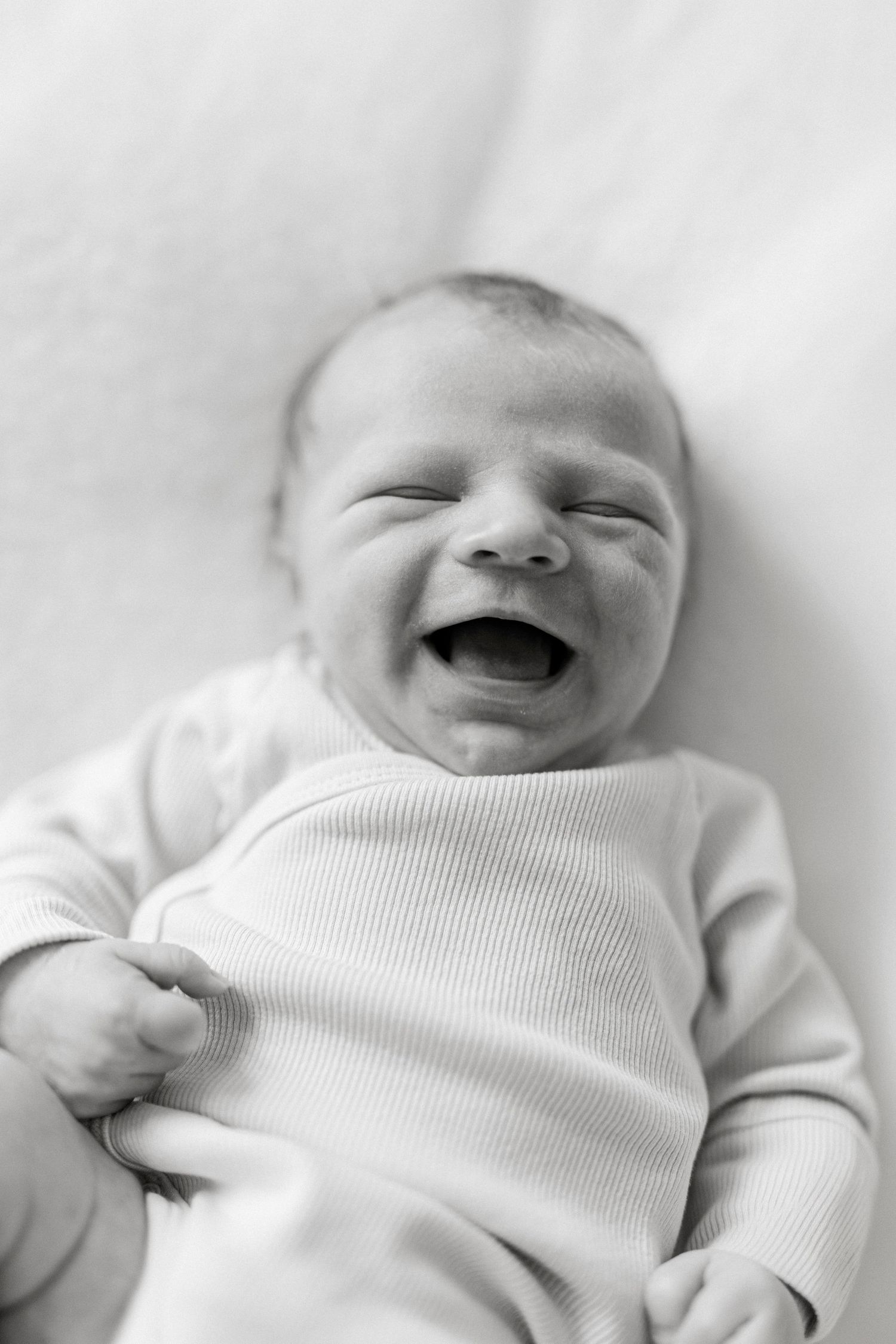 Black and white photo of a laughing newborn baby lying on a white blanket wearing a ribbed onesie.
