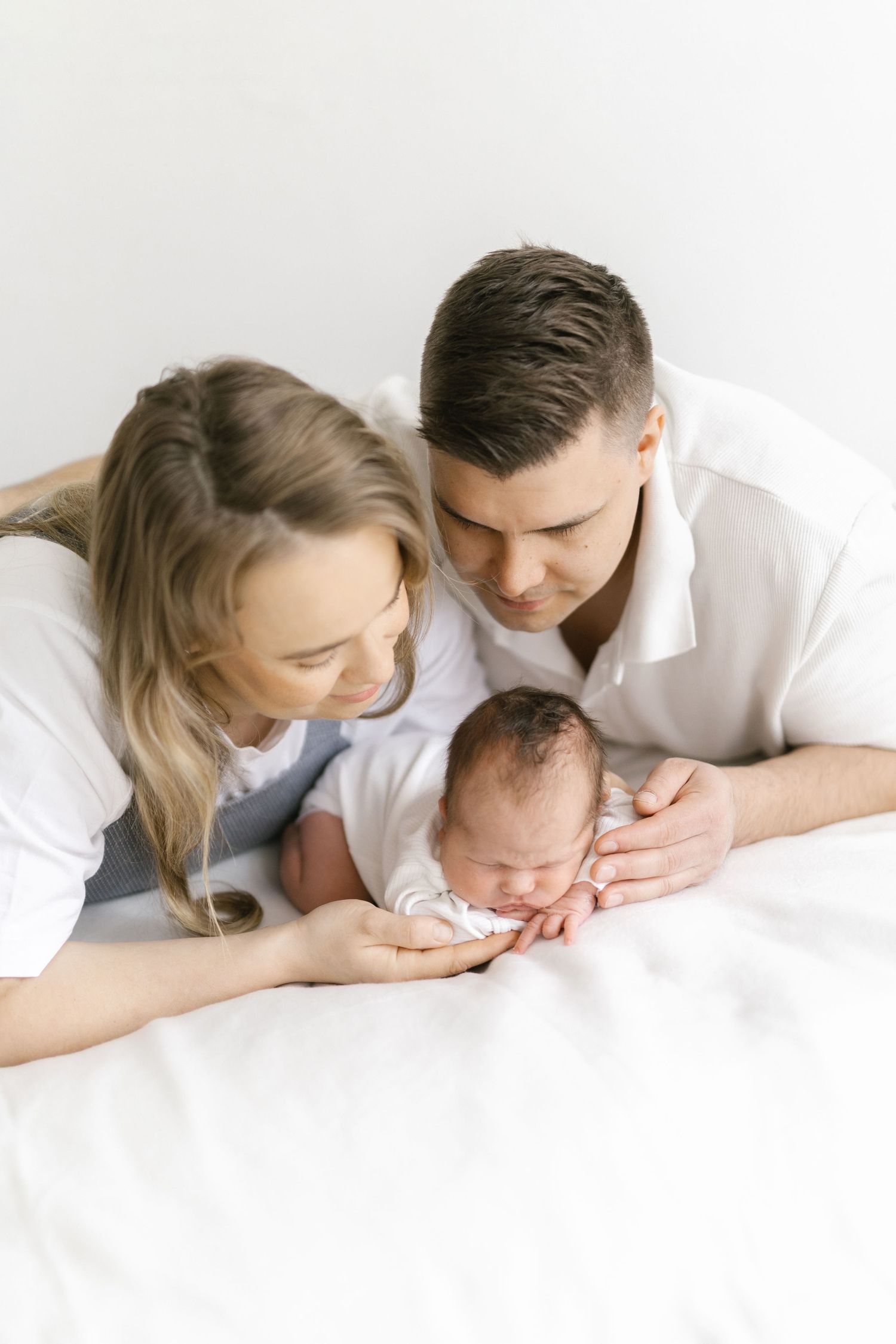A tender family moment captured in white as parents lean close to their sleeping newborn on a bright background.