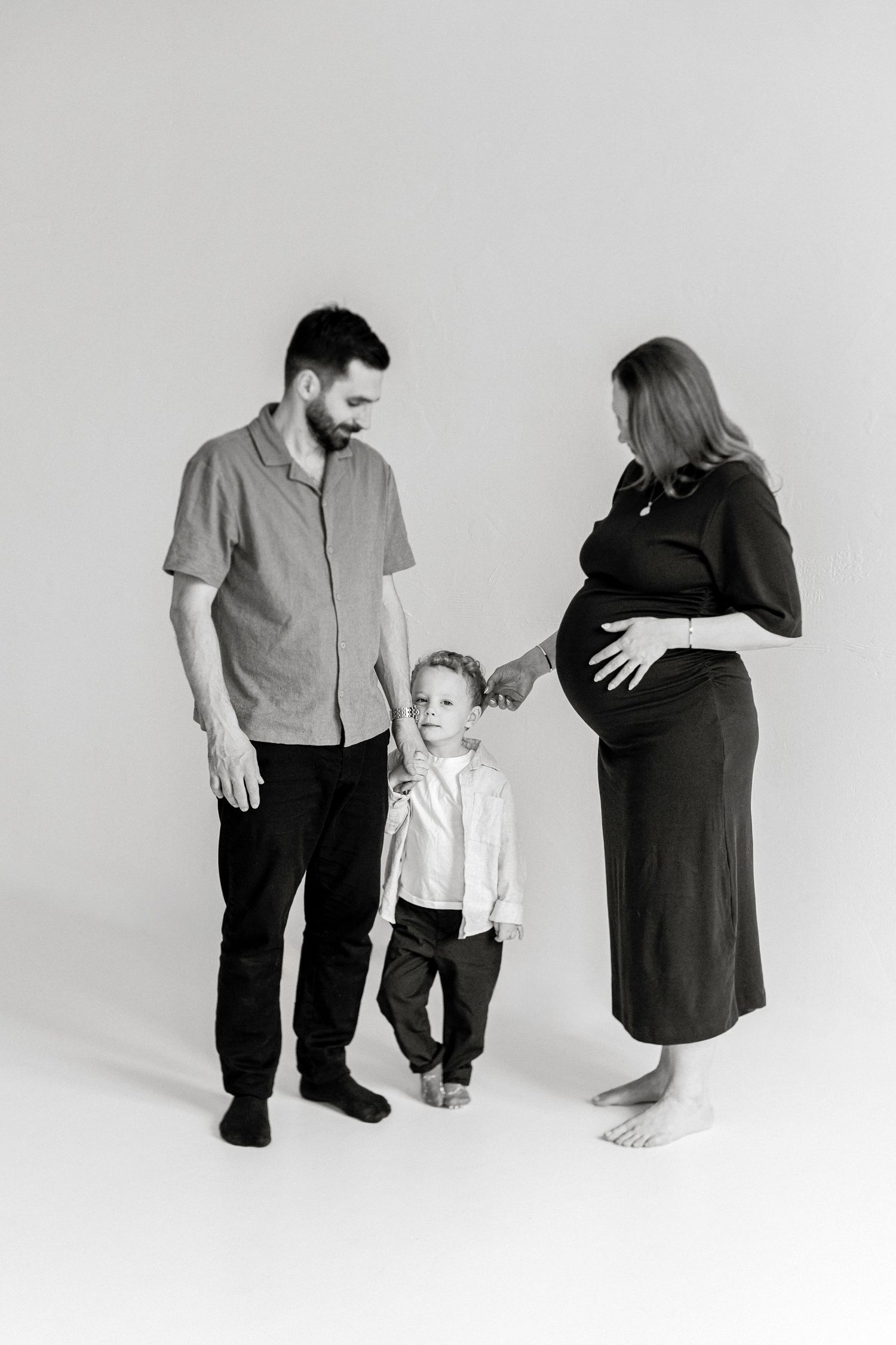 Black and white maternity photo of pregnant woman, bearded man, and young child posing together in a studio.