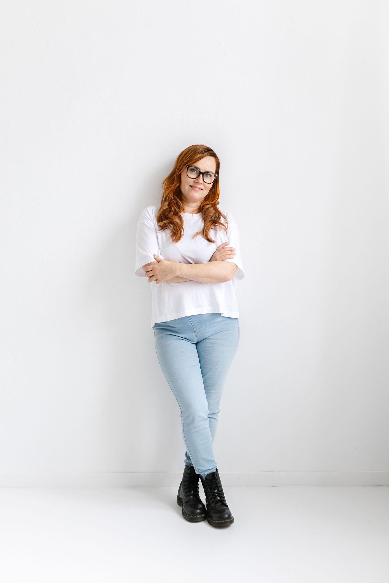 A person wearing glasses, a white t-shirt and light jeans stands with crossed arms against a white wall.