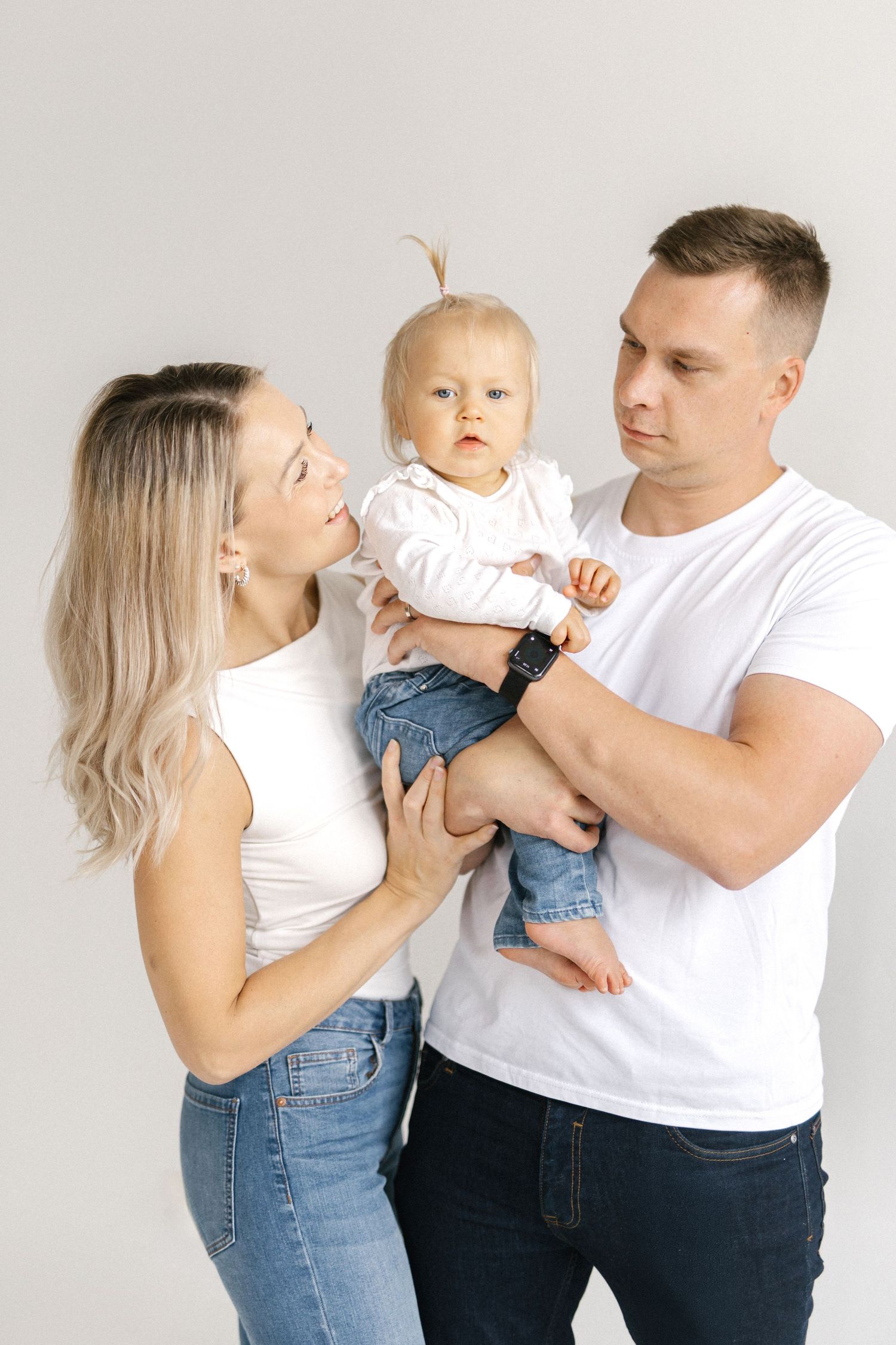 Young family in white tops and jeans posing with baby in studio, parents holding infant against white background.