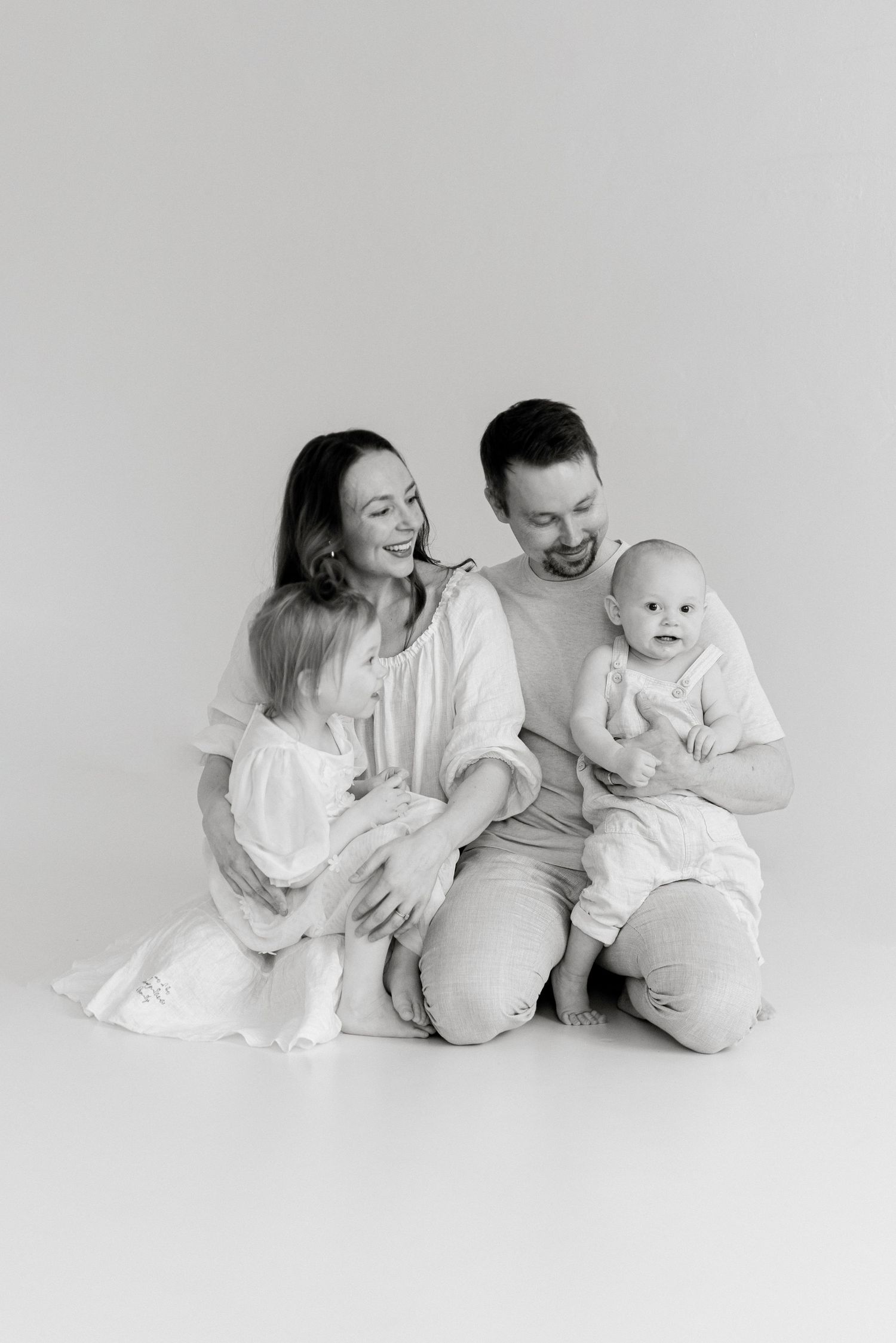 Black and white family portrait of parents sitting on floor with toddler girl and baby boy in white outfits.
