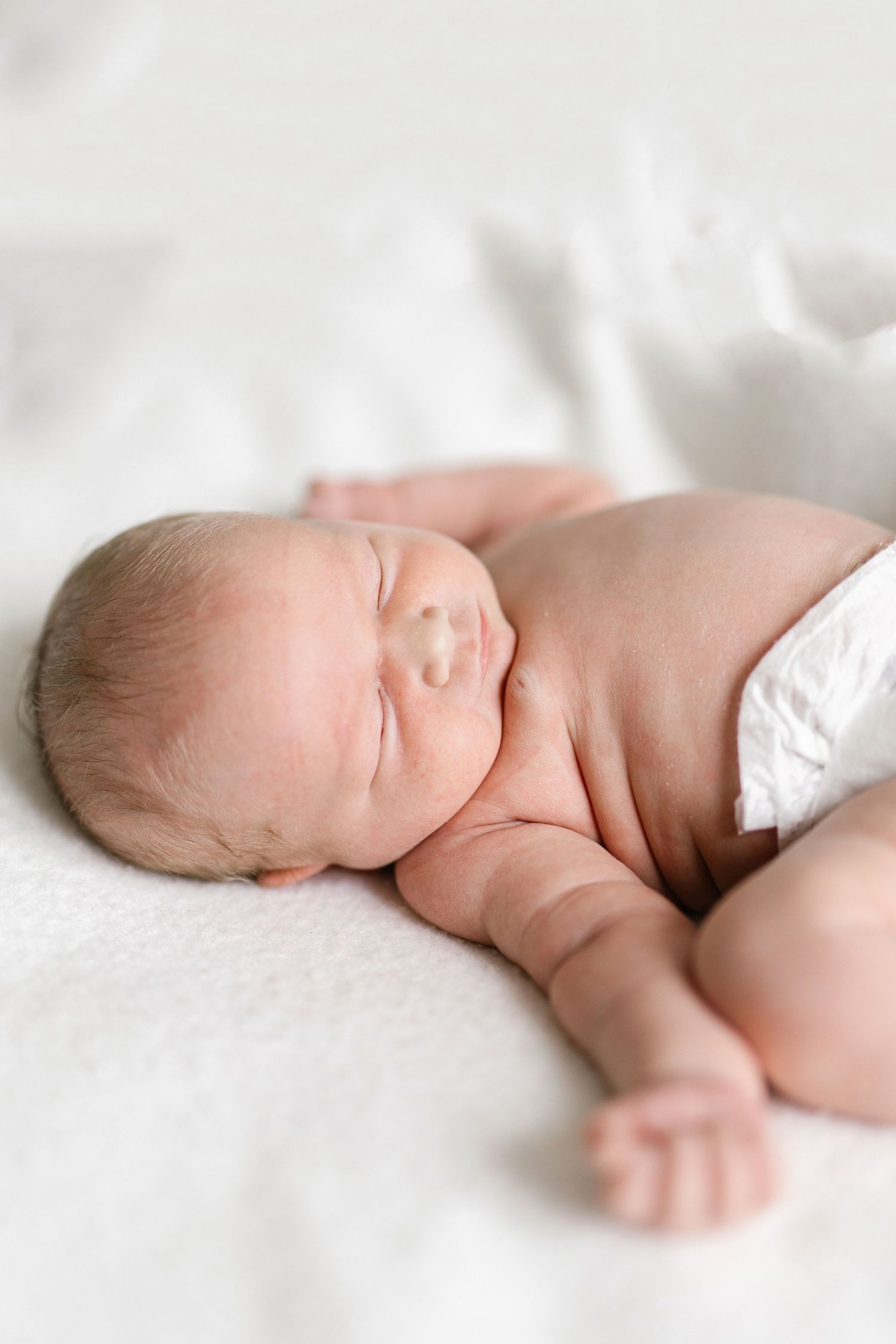 Peaceful newborn baby sleeping on a soft white blanket, wearing a diaper, with chubby cheeks and tiny hands visible.