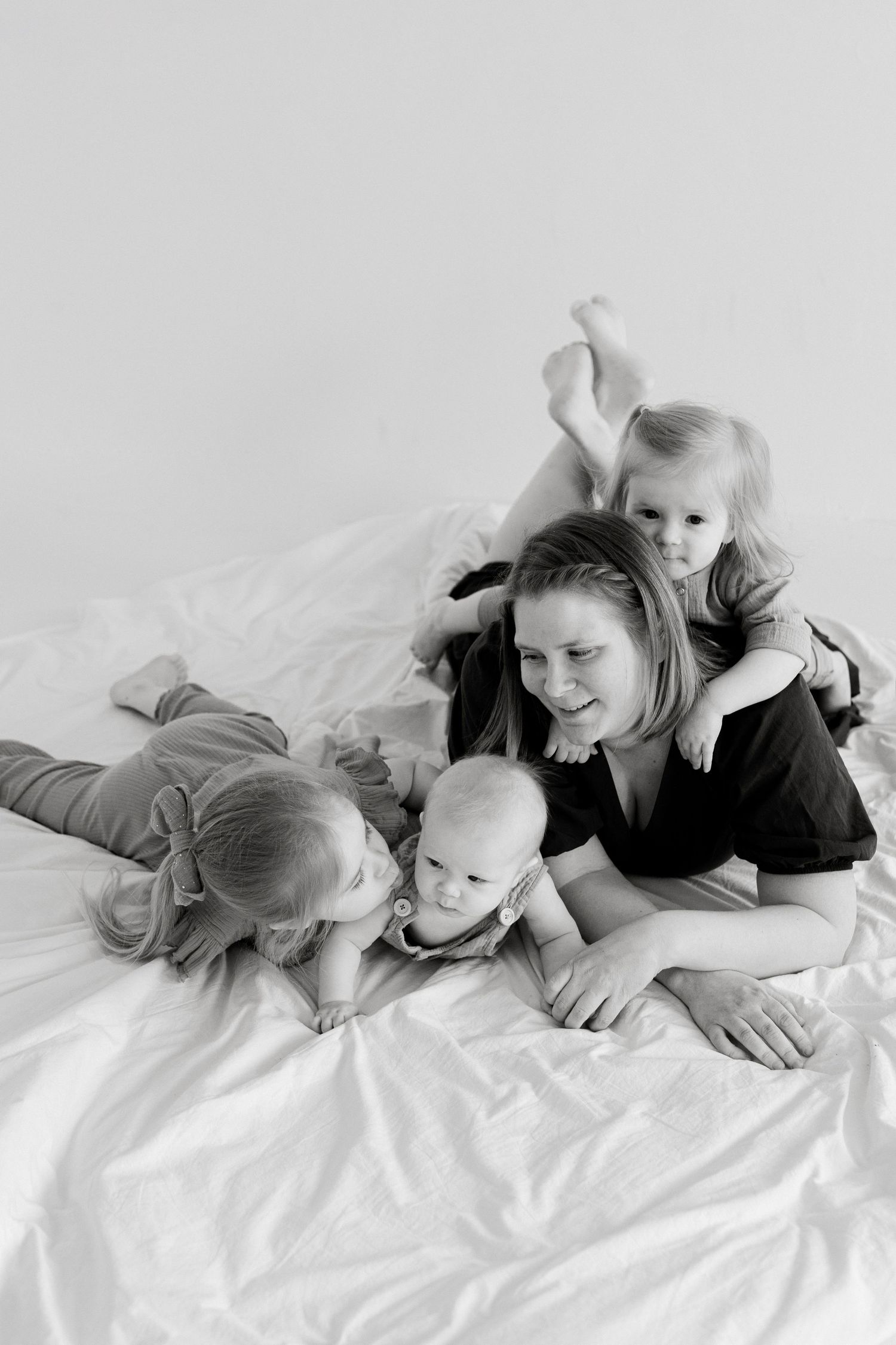Black and white photo of a mother lying on a bed with three young children, smiling and cuddling together.