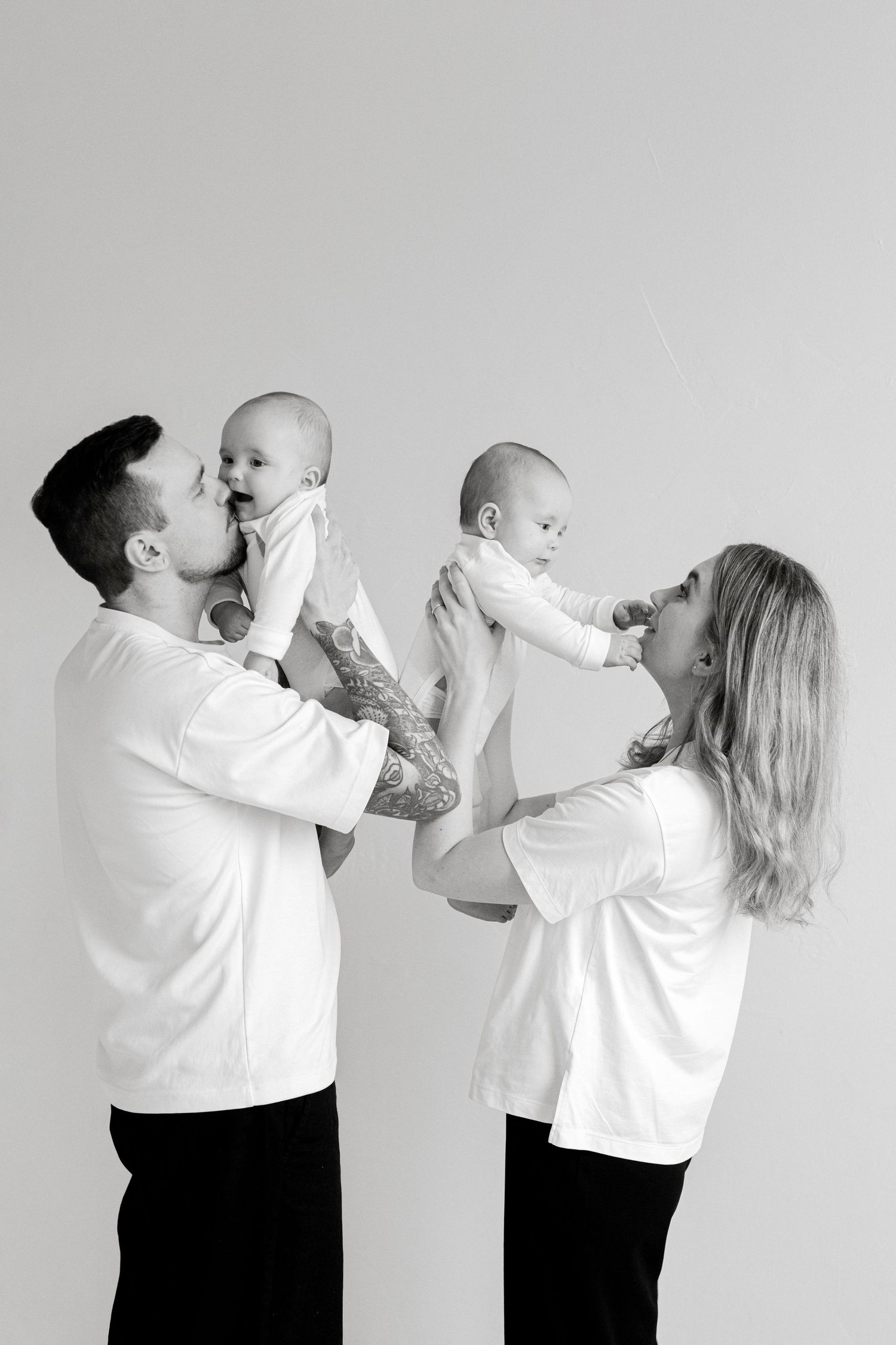 Black and white photo of parents in white shirts holding twin babies up in a studio portrait.