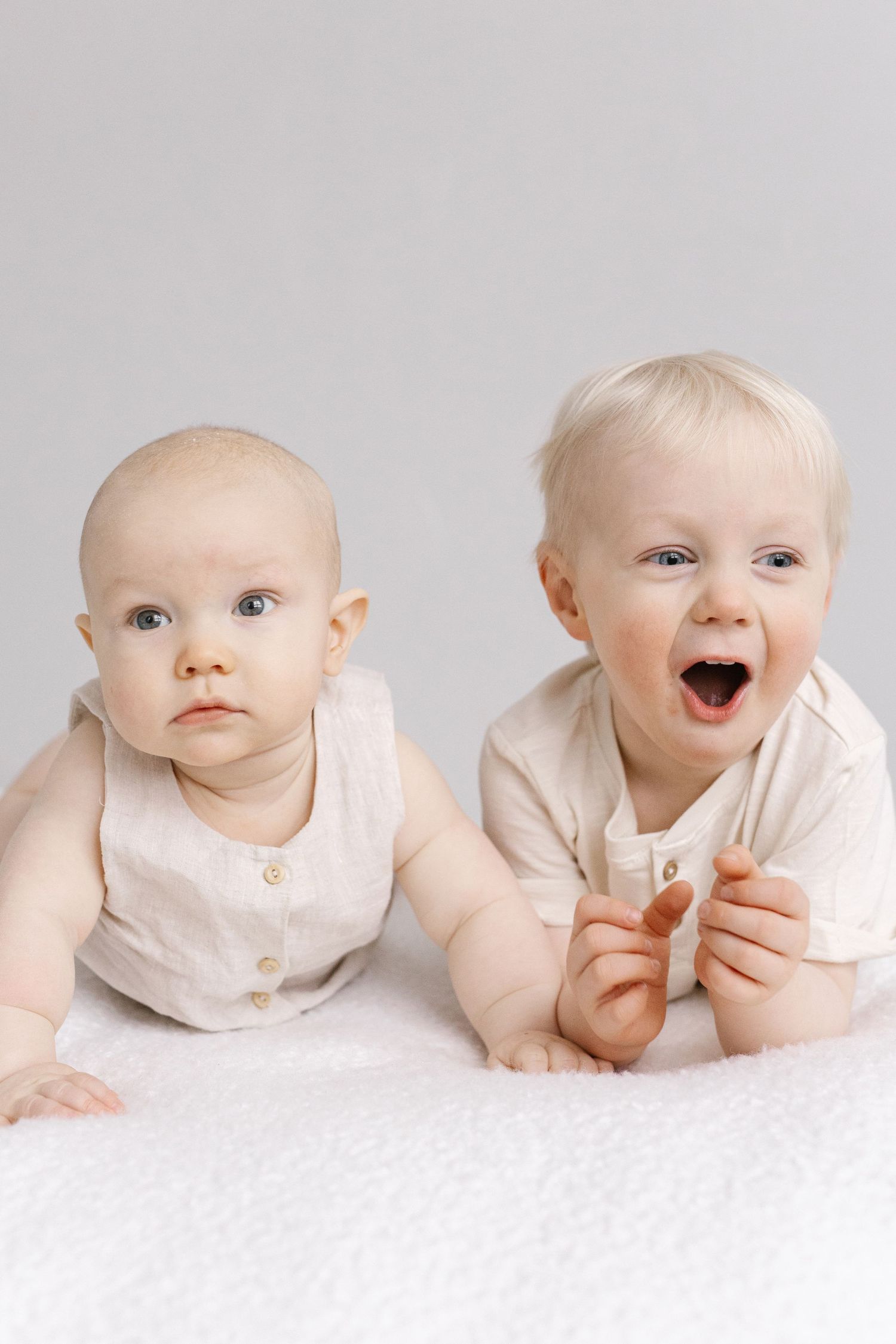Two adorable babies in cream linen outfits lying together, one smiling with mouth open.