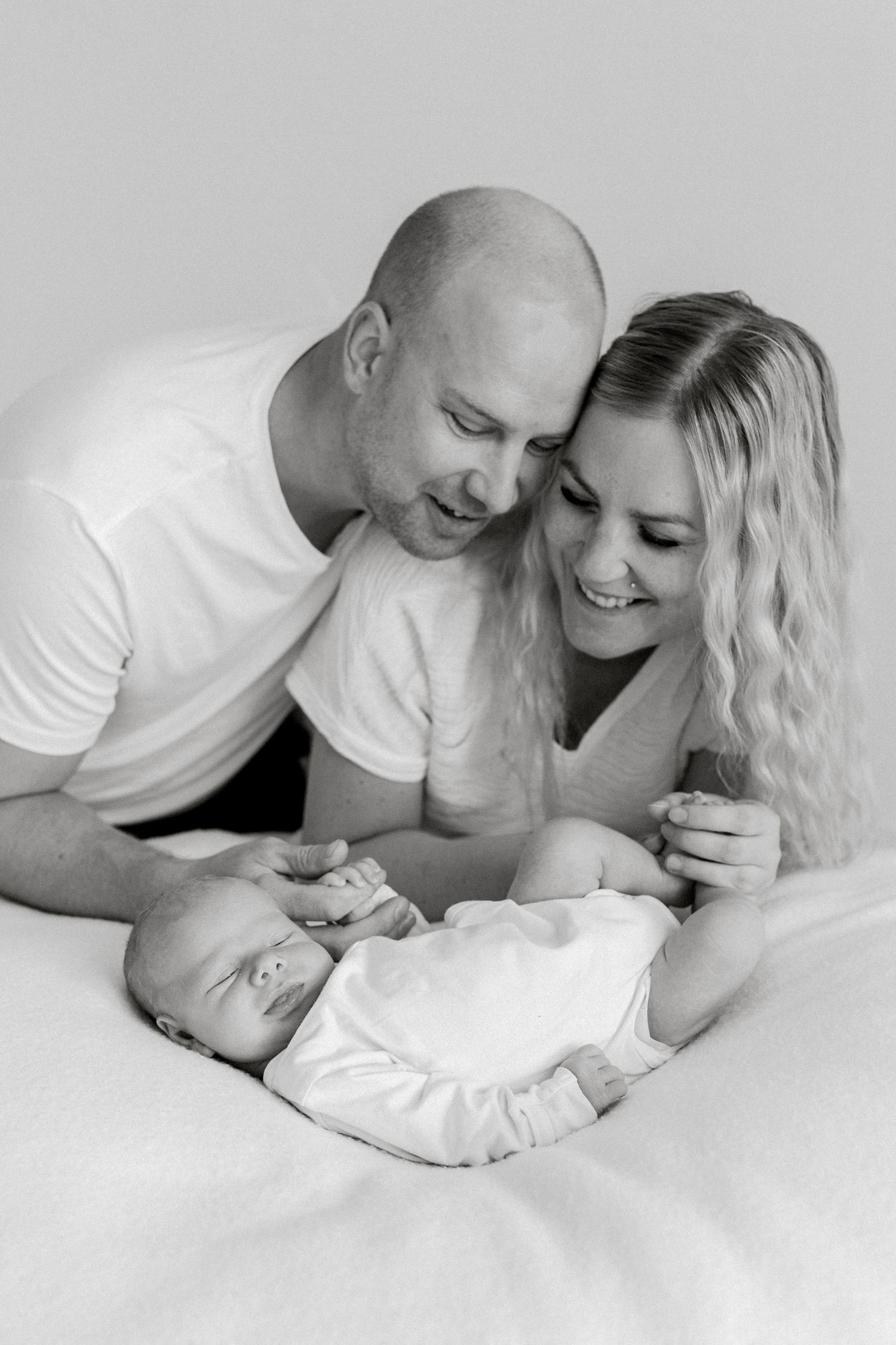 Black and white photo of smiling parents lovingly gazing at their newborn baby lying on a white bed.