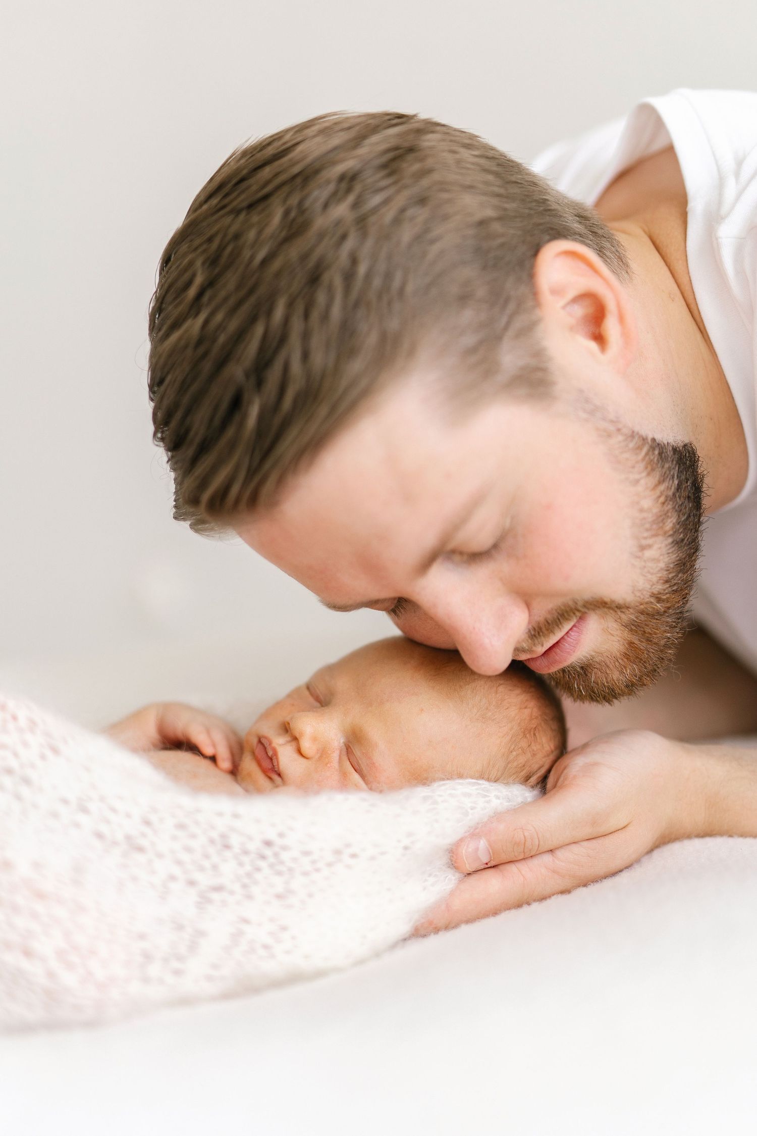A bearded father tenderly kisses his sleeping newborn baby wrapped in a soft pink blanket.