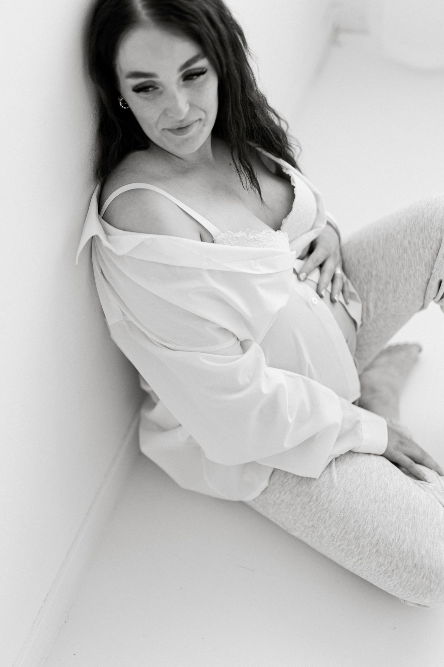 Black and white photo of a pregnant woman sitting against a wall, wearing an open white shirt, cradling her baby bump.