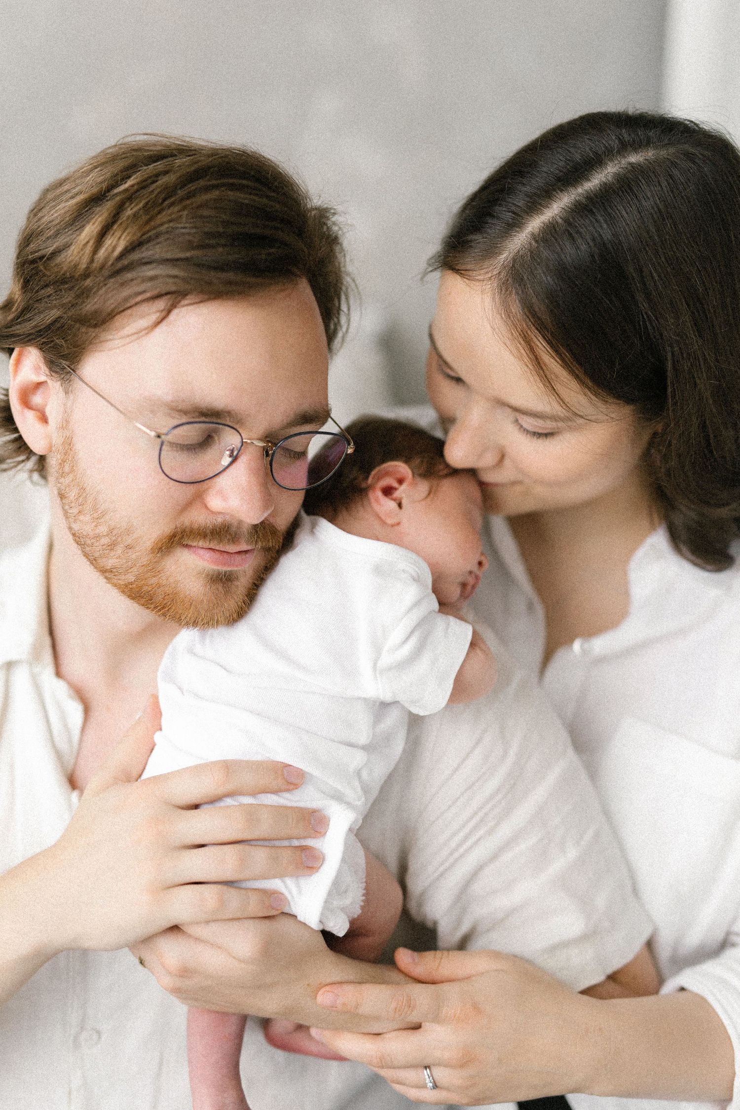 New parents tenderly holding their newborn baby, father with glasses and red beard, mother in white shirt.