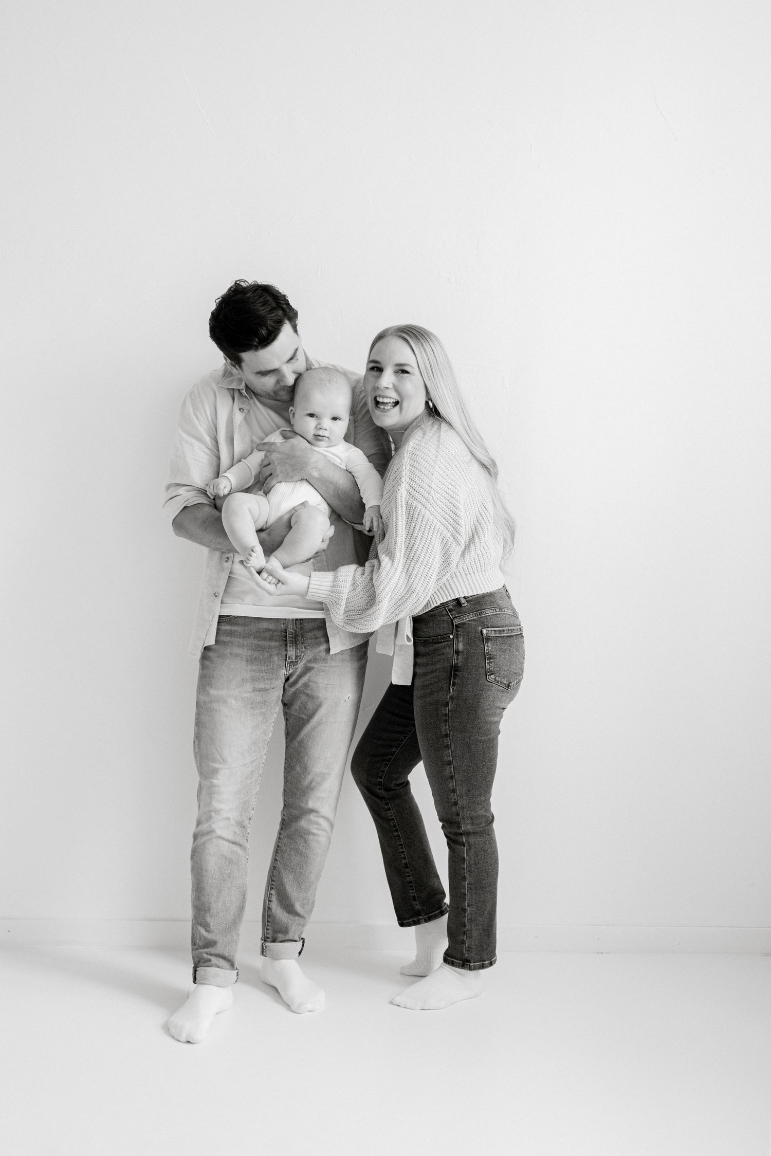 Black and white family portrait of parents holding their baby, smiling together against a white background.