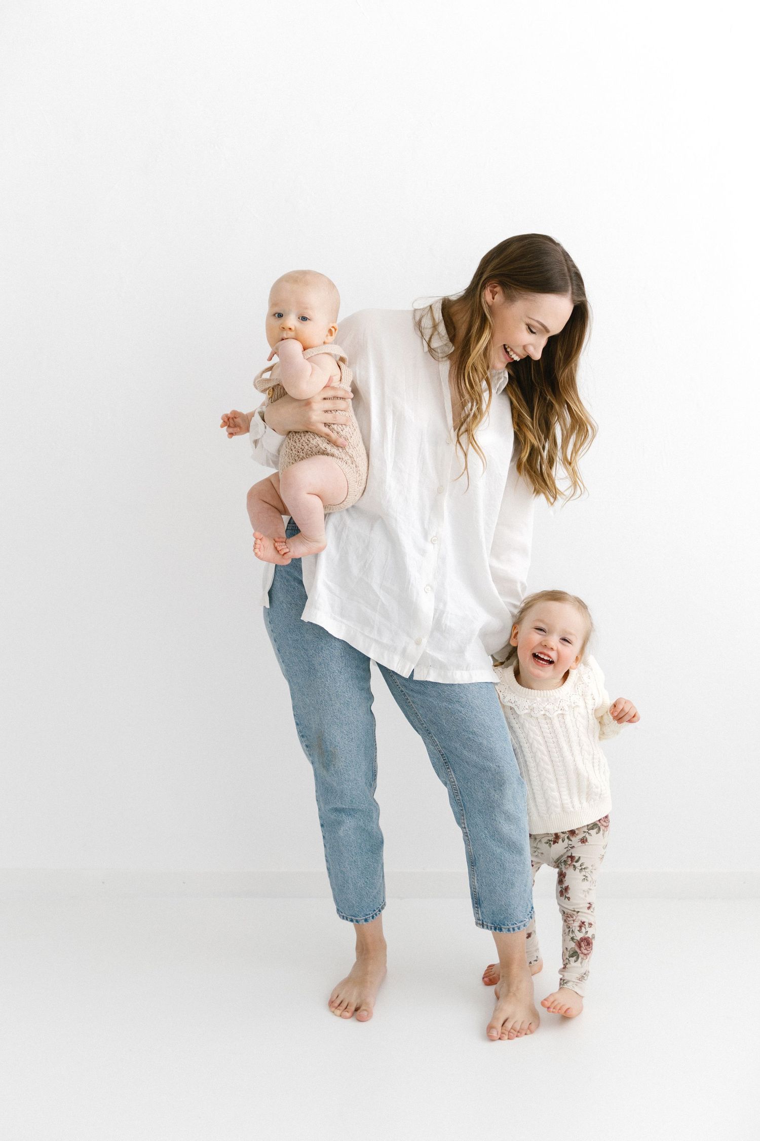 Smiling mother in white shirt holds baby while toddler daughter laughs beside her on white backdrop.
